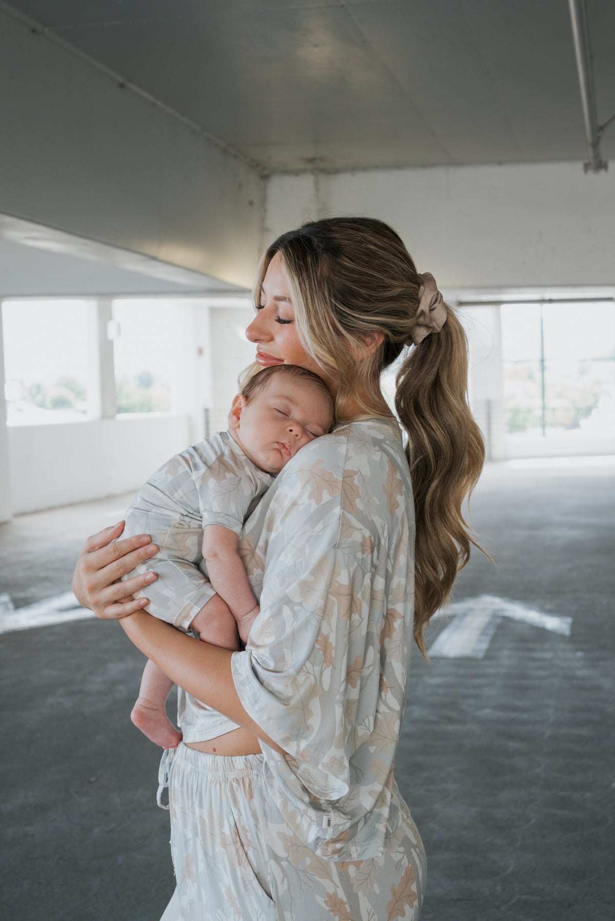 A woman with long hair in a ponytail holds a sleeping baby in her arms; both are wearing matching forever french baby Shortie Bamboo Zip pajamas in the Woodland print, standing together in a bright, empty parking garage.
