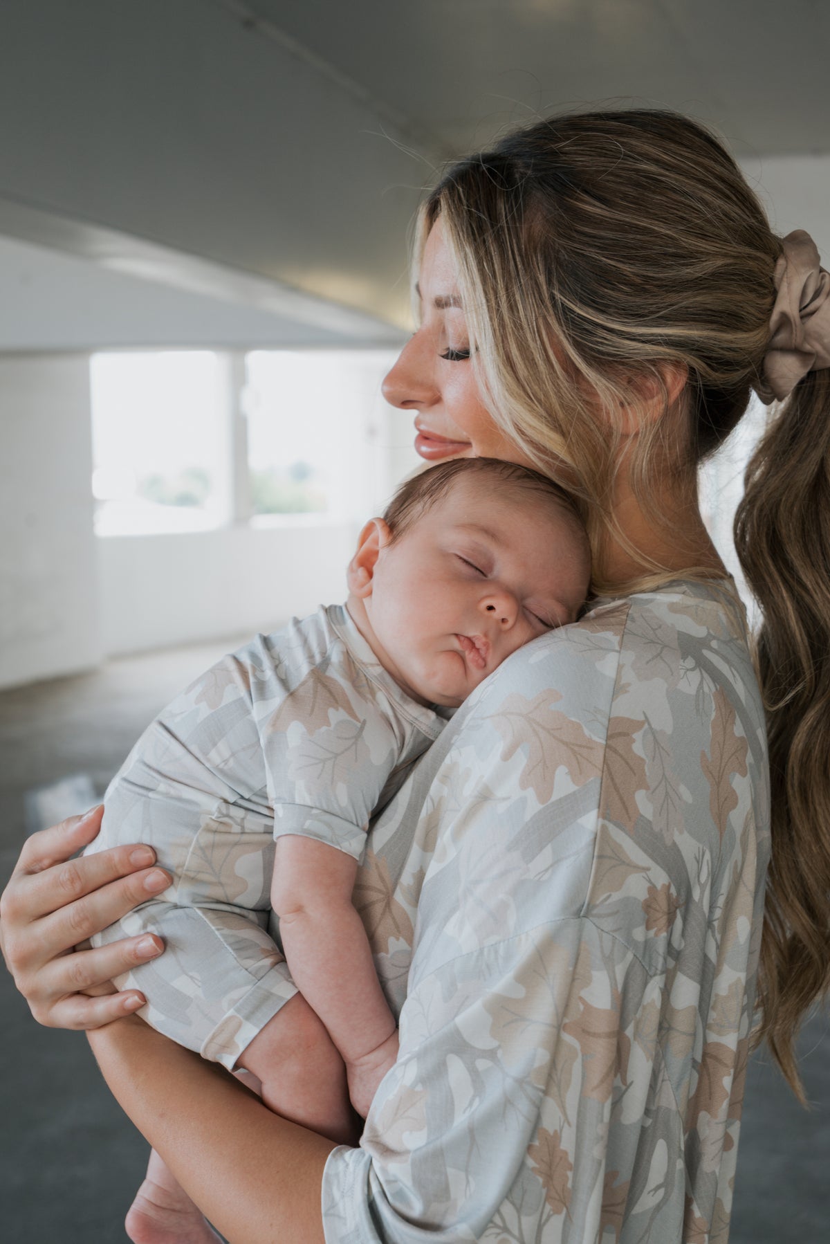 A woman with long, light brown hair pulled back in a scrunchie cradles a sleeping baby against her chest. Both are indoors in a softly lit space, wearing matching, light-colored leaf-patterned outfits from forever french baby—the little one snuggled in the cozy Shortie Bamboo Zip | Woodland romper.
