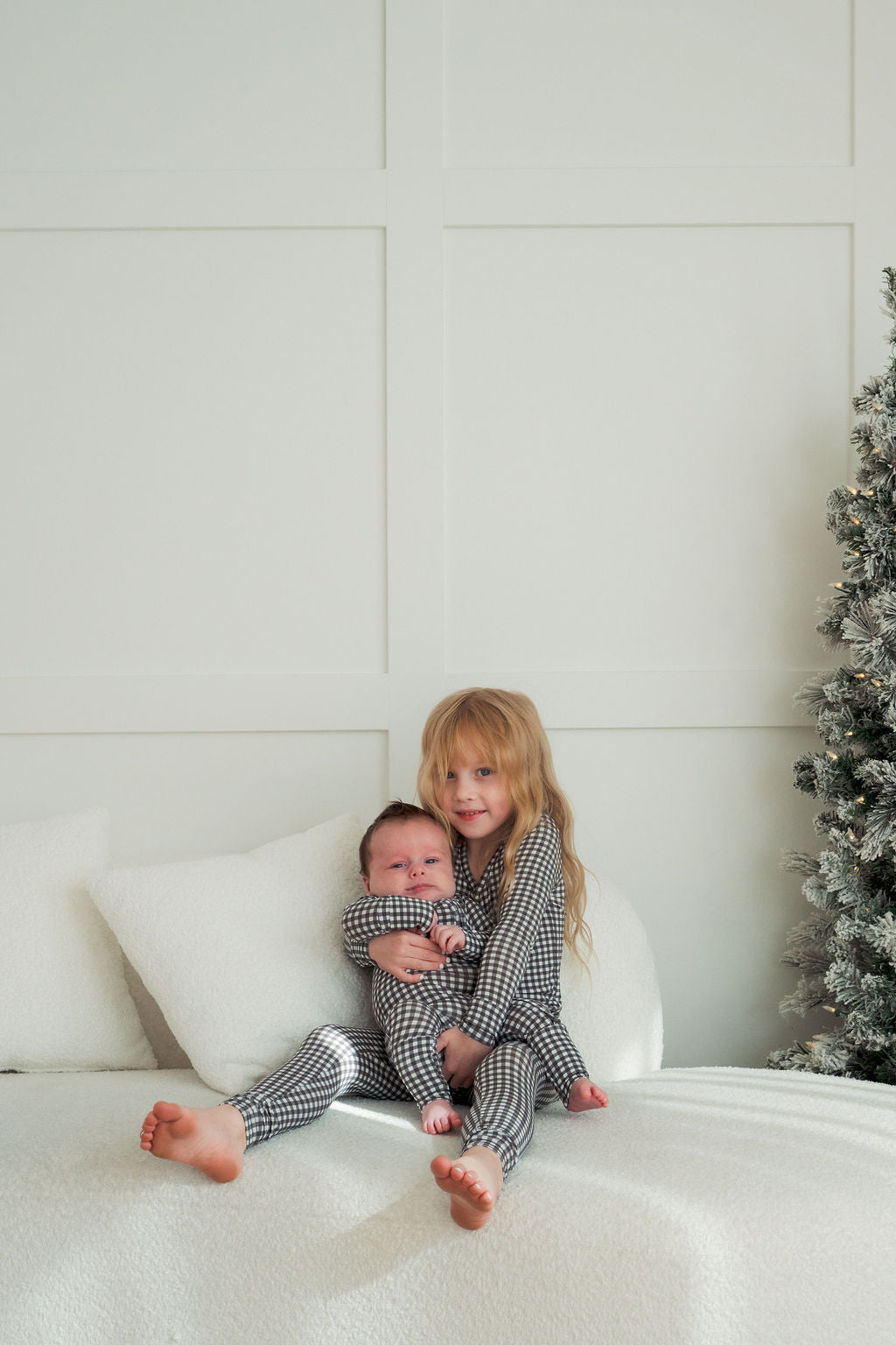 Two young children wear matching forever french baby Bamboo Two Piece Pajamas in Green Gingham as they sit on a bed. The older hugs the younger on their lap, next to a pillow and Christmas tree, for a cozy holiday scene.
