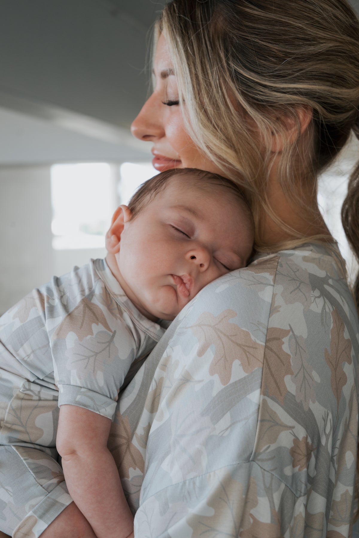 A woman smiles softly while holding a sleeping baby on her shoulder. Both are wearing matching, light-colored pajamas featuring the woodland leaf print from forever french baby. The baby rests peacefully in the cozy Shortie Bamboo Zip | Woodland romper, and the woman looks content.