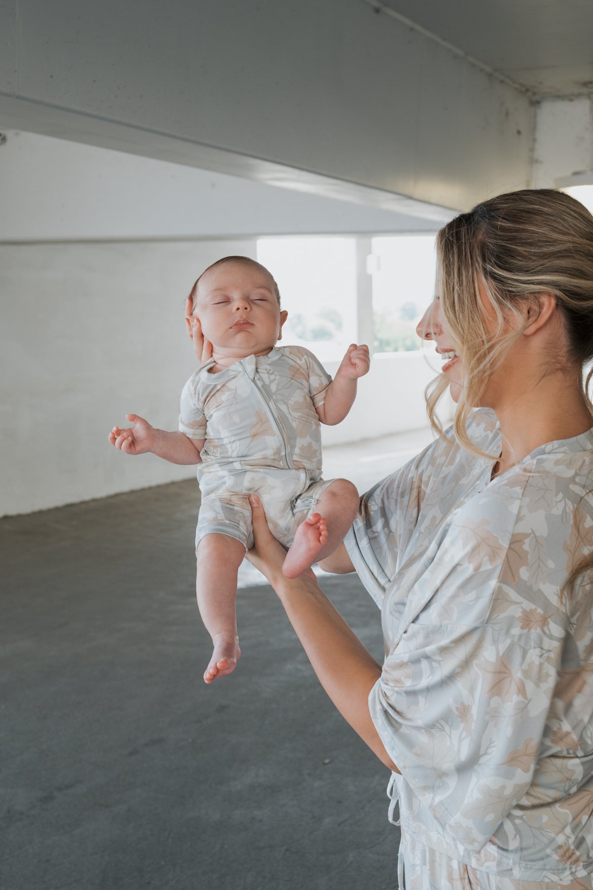 A woman with blonde hair holds a baby dressed in the forever french baby Shortie Bamboo Zip | Woodland romper—a soft, hypoallergenic piece of sleepwear featuring a charming woodland print—in a bright, spacious indoor setting. Both wear light, patterned outfits as the baby rests comfortably with closed eyes and relaxed arms.