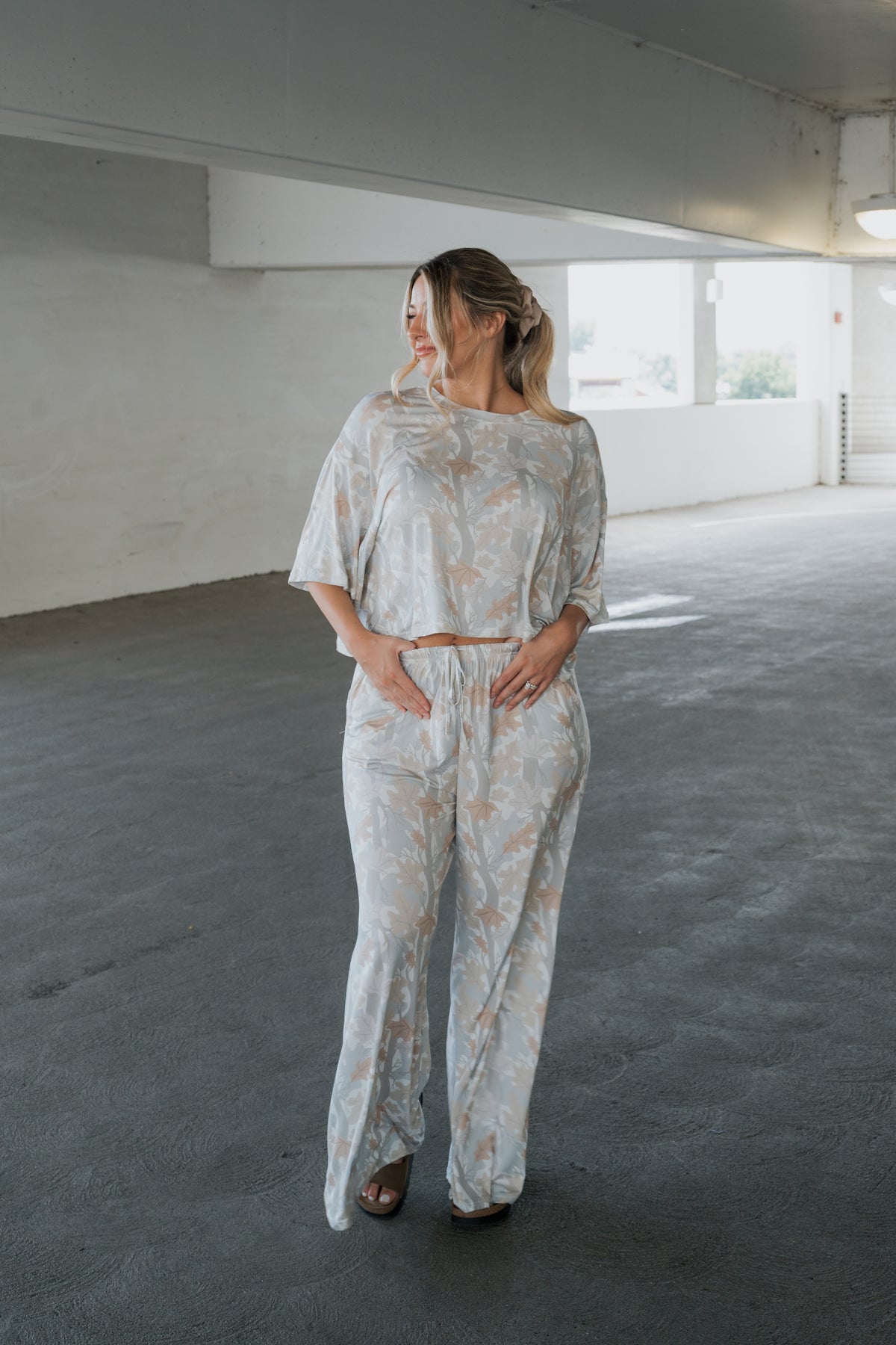 A woman stands in a parking garage wearing the forever french baby Women’s Bamboo Crop Tee Lounge Set in Woodland—a light, loose-fitting matching set featuring a crop tee and wide-leg pants with a subtle leaf pattern. She faces left with her hands in her pockets, blonde hair styled in a ponytail.