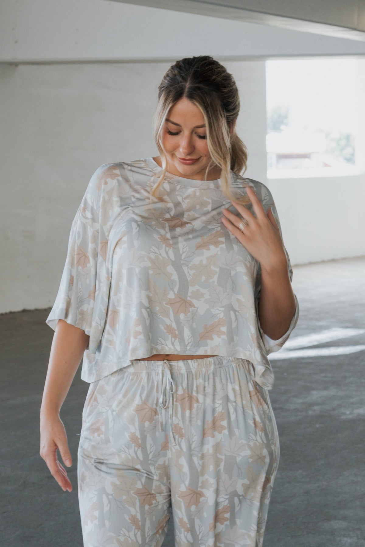 A woman in a loose, light-colored, leaf-patterned matching top and pants stands indoors, looking down and touching her hair. Her Women’s Boxy Crop Tee | Woodland from forever french baby not only offers comfort and style but also elevates her look with its natural, airy fabric.
