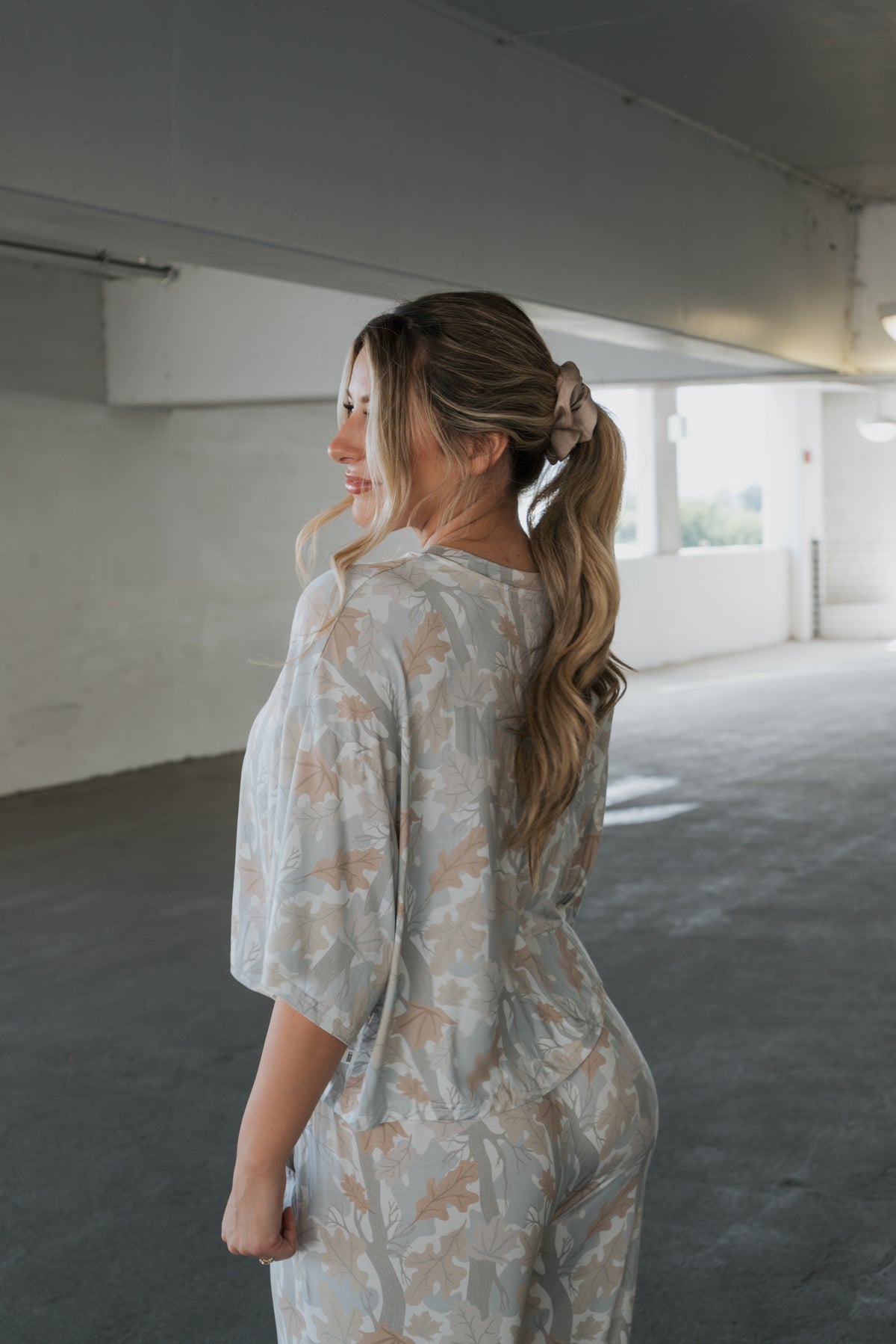 A woman with long, wavy hair in a ponytail smiles and looks to her left as she stands in a parking garage wearing the forever french baby Women’s Bamboo Crop Tee Lounge Set in Woodland.
