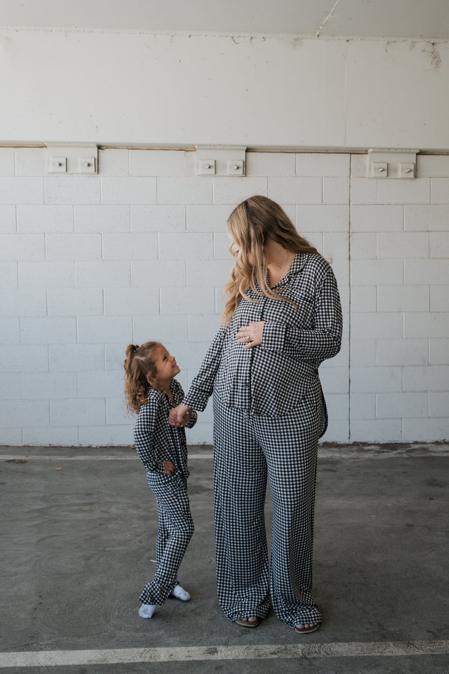 A woman and a young girl, both wearing matching forever french baby Black Gingham bamboo outfits—including the Girl’s Bamboo Button Up Lounge Set—stand together in a parking garage, holding hands and gazing at each other.