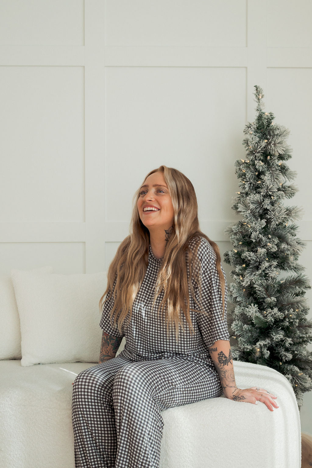 A woman with long hair and tattoos, wearing the forever french baby Women's Boxy Tee Lounge Set in Green Gingham, smiles while sitting on a white sofa near a small frosted artificial Christmas tree in a bright room.