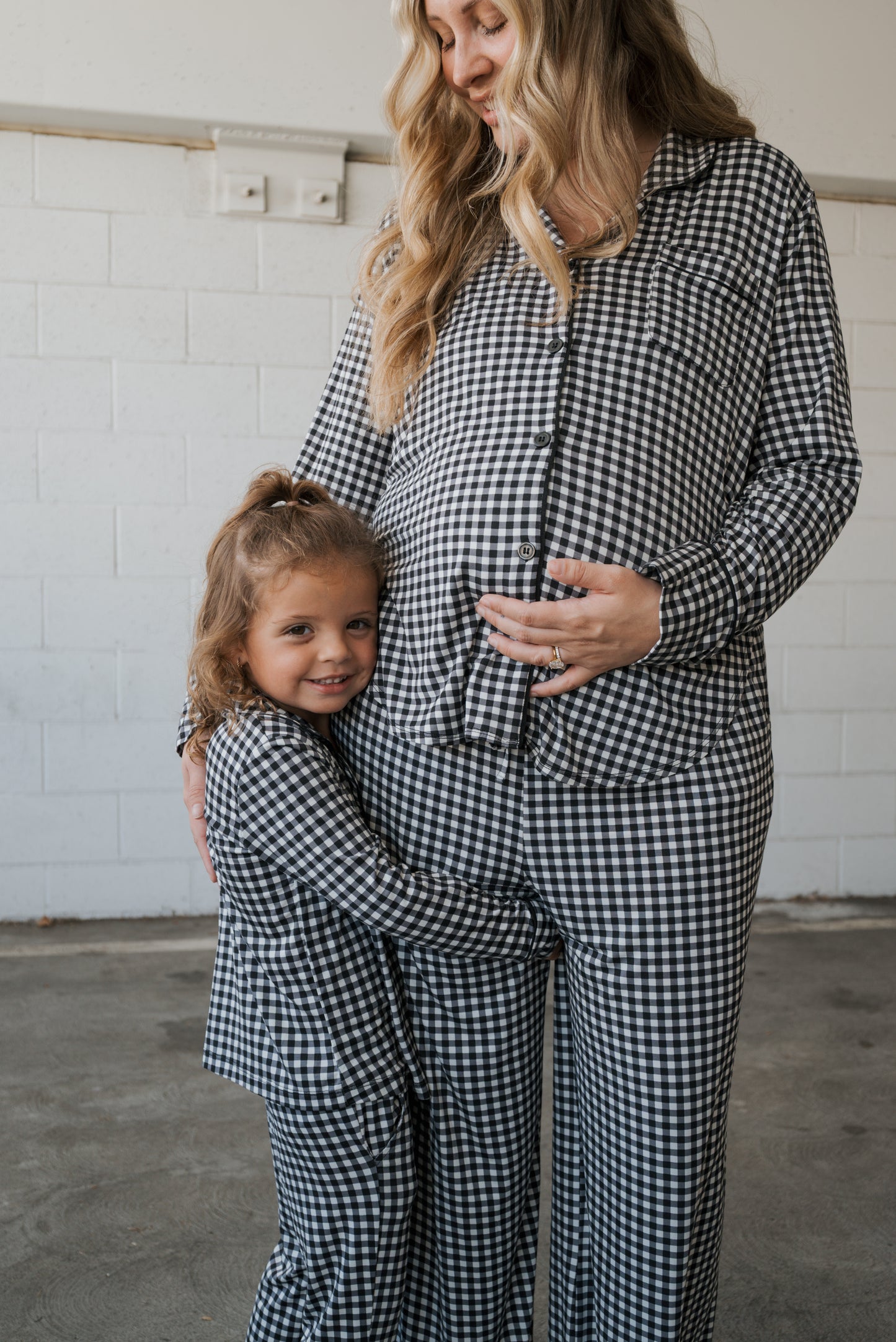 A young girl hugs a pregnant woman’s belly as they both smile, wearing matching forever french baby Girl’s Bamboo Button Up Lounge Sets in Black Gingham, standing in front of a white brick wall.
