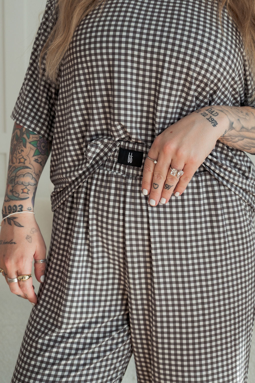 A person with tattooed arms and rings wears the forever french baby Women's Boxy Tee Lounge Set in green gingham, holding part of the loose-fitting top as it drapes over the matching pants.