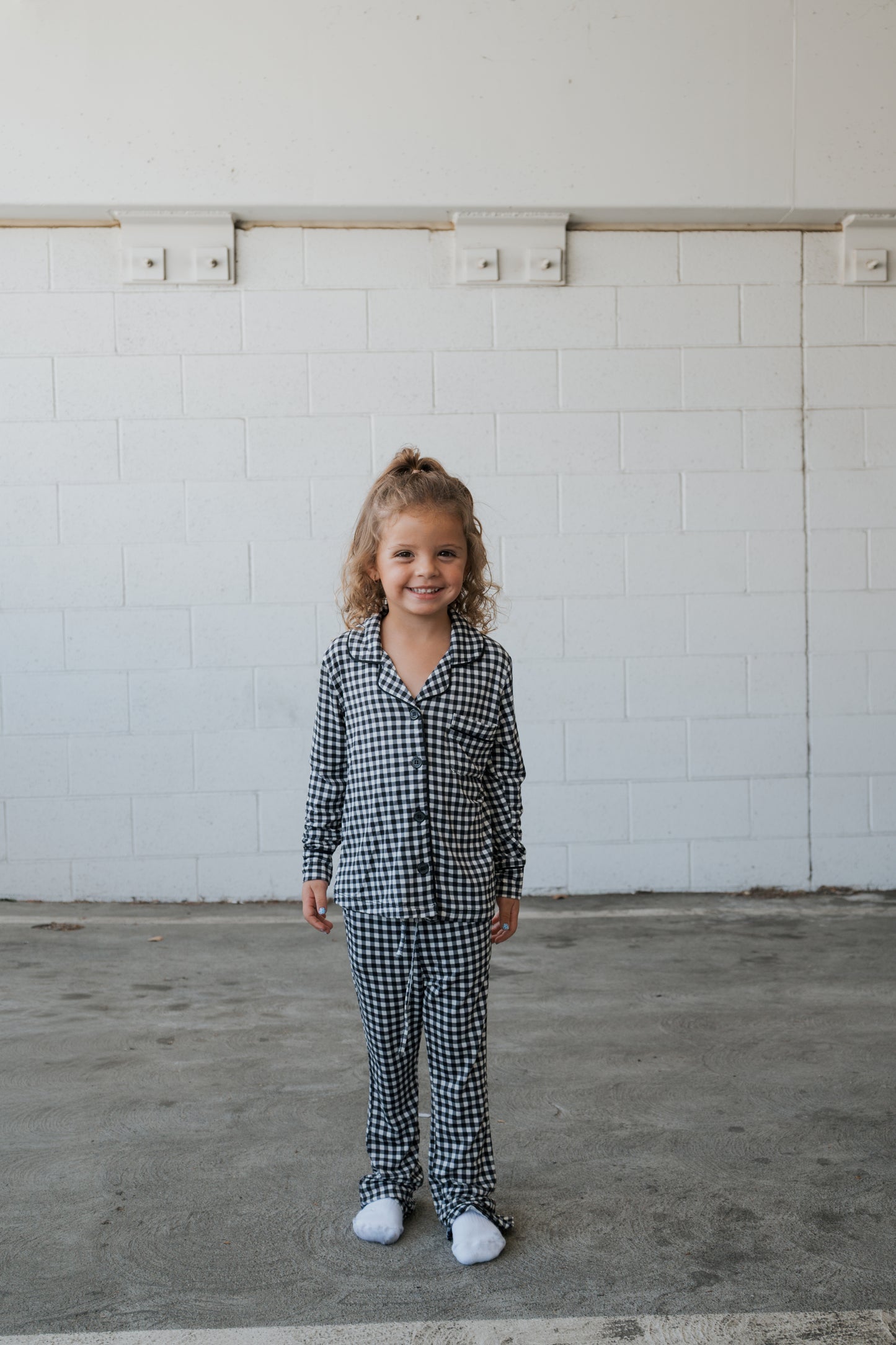 A young child with curly hair stands smiling, wearing the forever french baby Girl’s Bamboo Button Up Lounge Set in Black Gingham with white socks, against a white brick wall background.