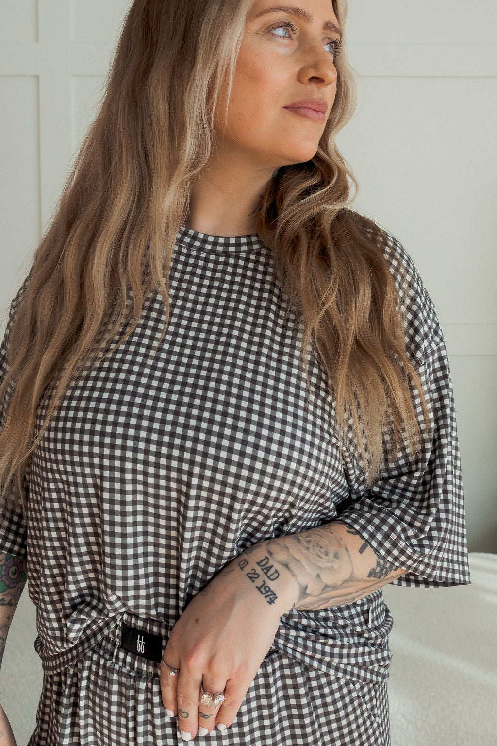 A woman with long, wavy blonde hair and visible forearm tattoos stands confidently against a white background, wearing the forever french baby Women's Boxy Tee Lounge Set in Green Gingham.