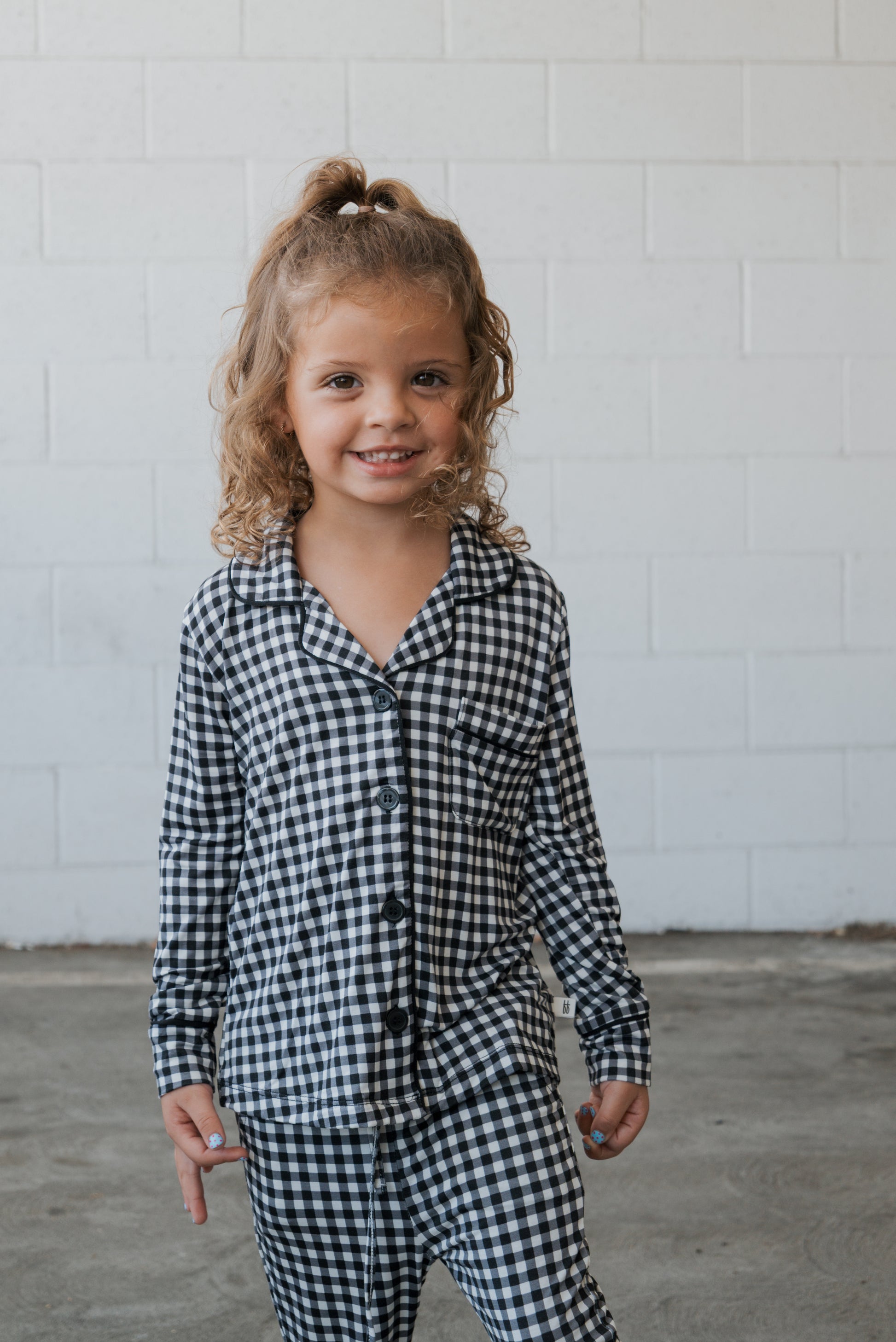 A young child with curly hair smiles while wearing the forever french baby Girl’s Bamboo Button Up Lounge Set in Black Gingham, standing in front of a white brick wall.