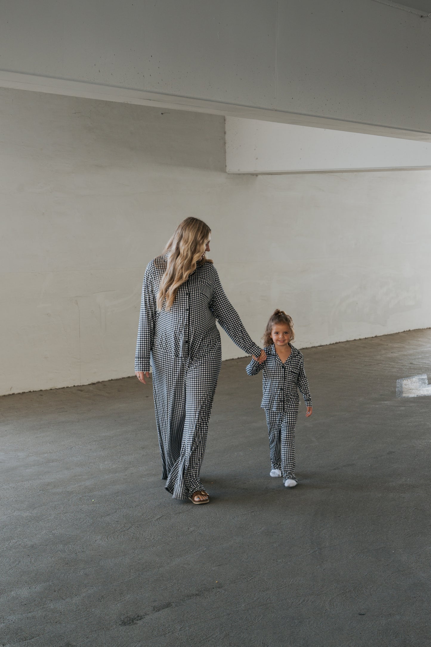 A woman and a young girl walk hand in hand through a spacious, empty indoor area with white walls, both dressed in forever french baby’s matching bamboo lounge sets. The young girl wears the Girl’s Bamboo Button Up Lounge Set in Black Gingham, their comfortable checkered clothing perfectly highlighting their special bond.