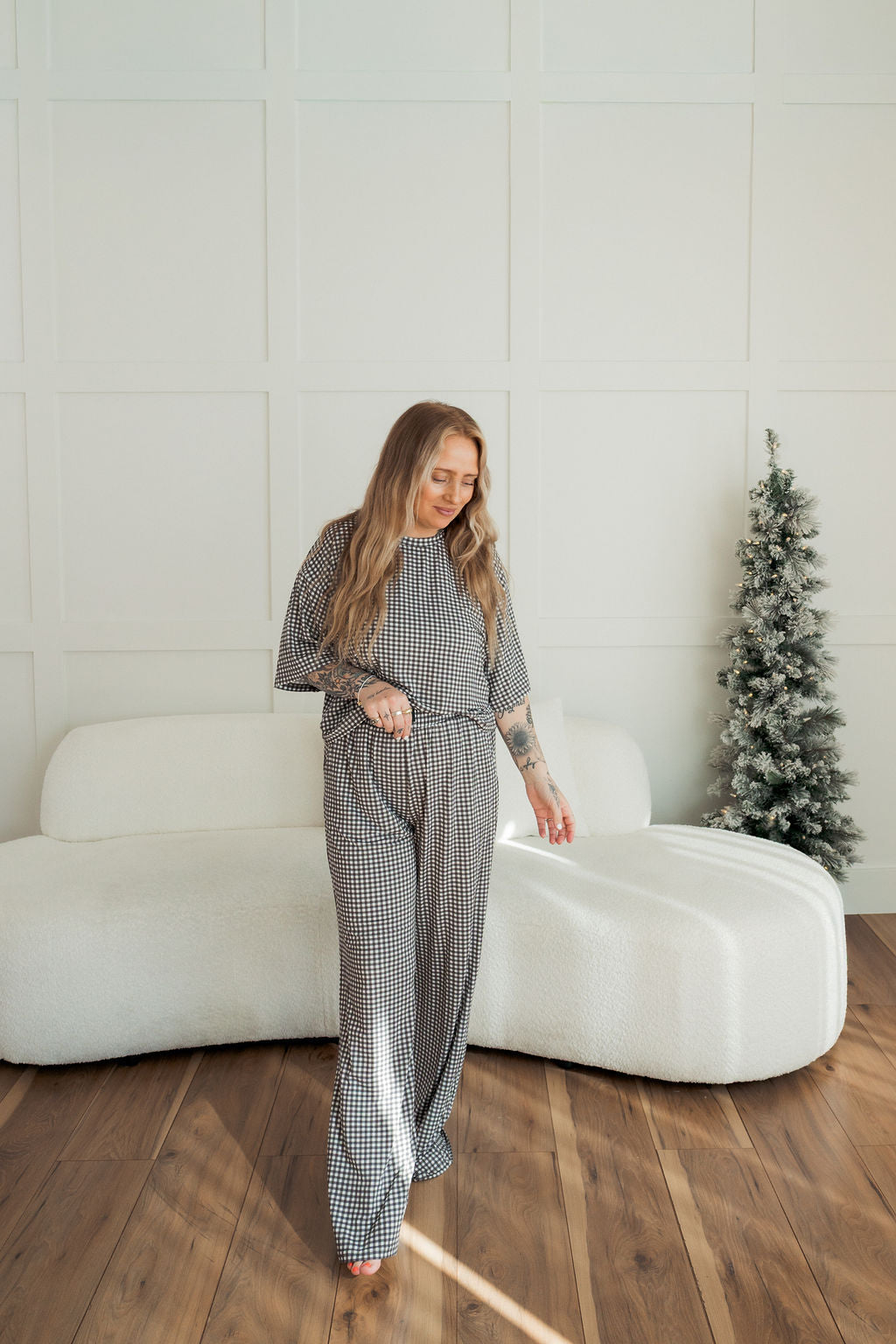 A woman with long, wavy hair smiles in front of a modern sofa and Christmas tree, wearing the forever french baby Women's Boxy Tee Lounge Set in Green Gingham, lit by natural light on a wooden floor.