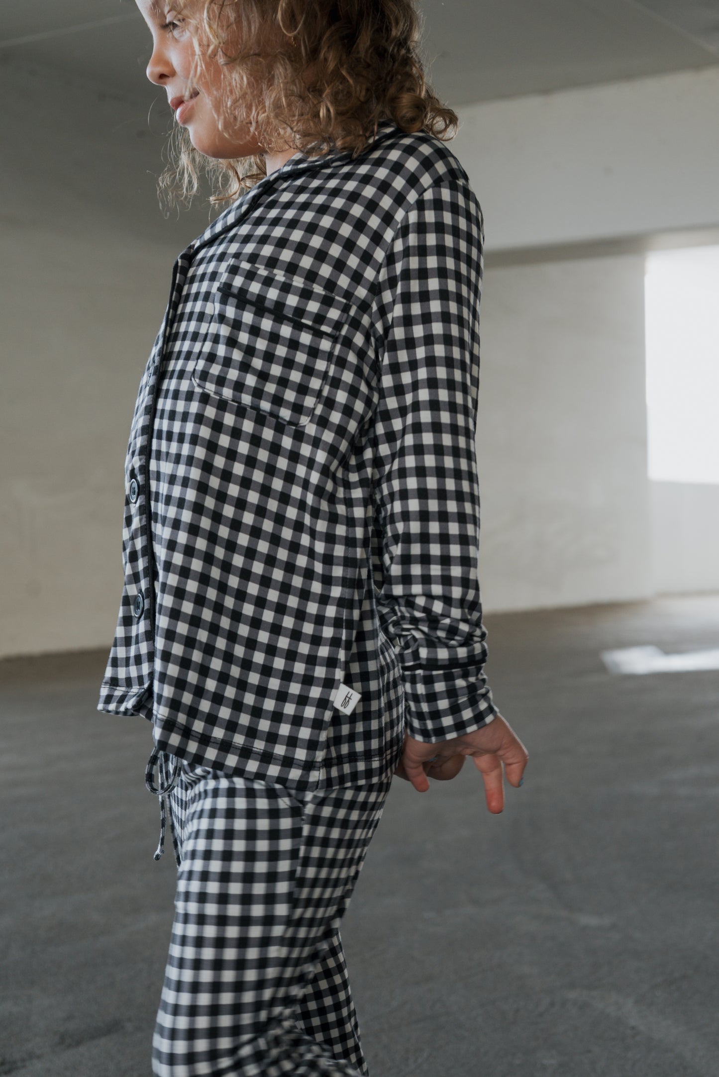 A child stands indoors in a minimal, sunlit room, wearing the forever french baby Girl’s Bamboo Button Up Lounge Set in Black Gingham, with one hand behind their back and looking to the side.
