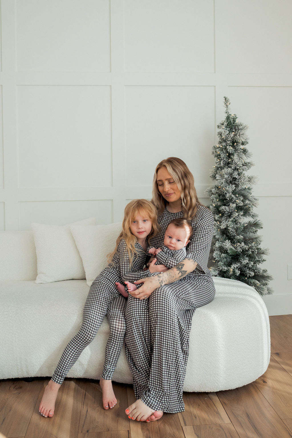 A woman and two children relax on a white sofa next to a frosted Christmas tree, all in forever french baby’s Bamboo Zip Pajamas in Green Gingham. The woman holds the baby while the older child leans against her, looking cozy.