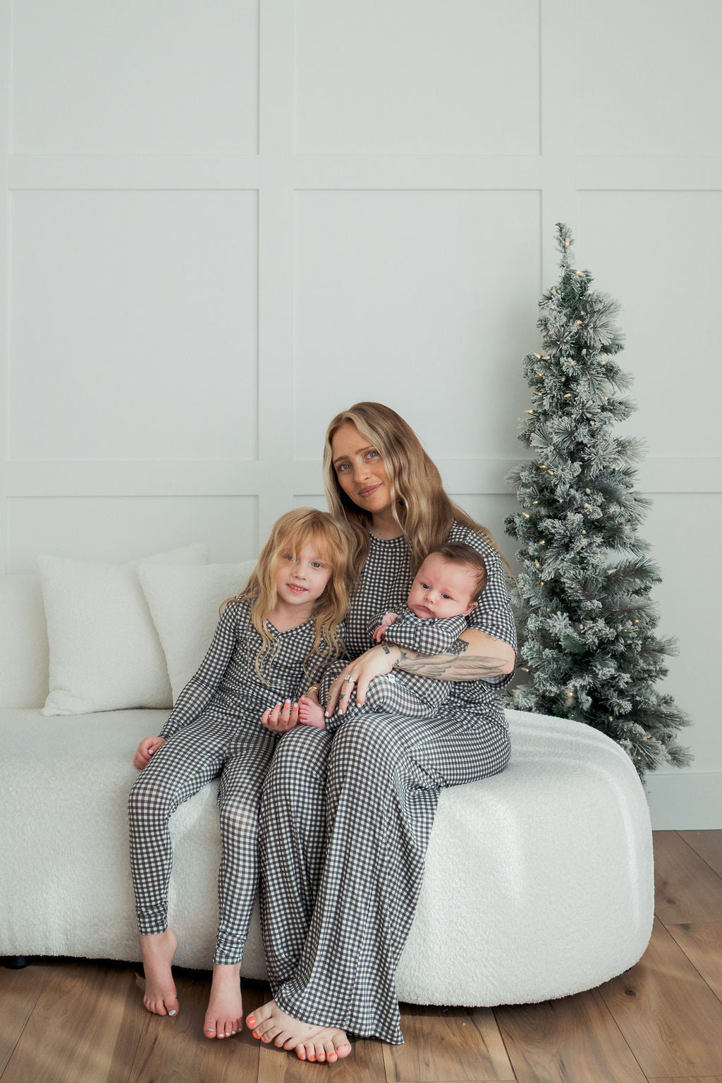 A woman sits on a white sofa holding a baby, with a young girl beside her. All three wear matching forever french baby Bamboo Two Piece Pajamas in green gingham. A frosted Christmas tree stands nearby against a white paneled wall.