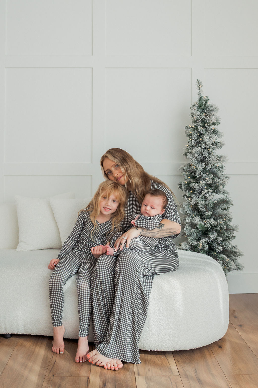 A woman and two children in matching forever french baby Women's Boxy Tee Lounge Set in Green Gingham sit on a white couch beside a small frosted Christmas tree in a bright, modern living room.