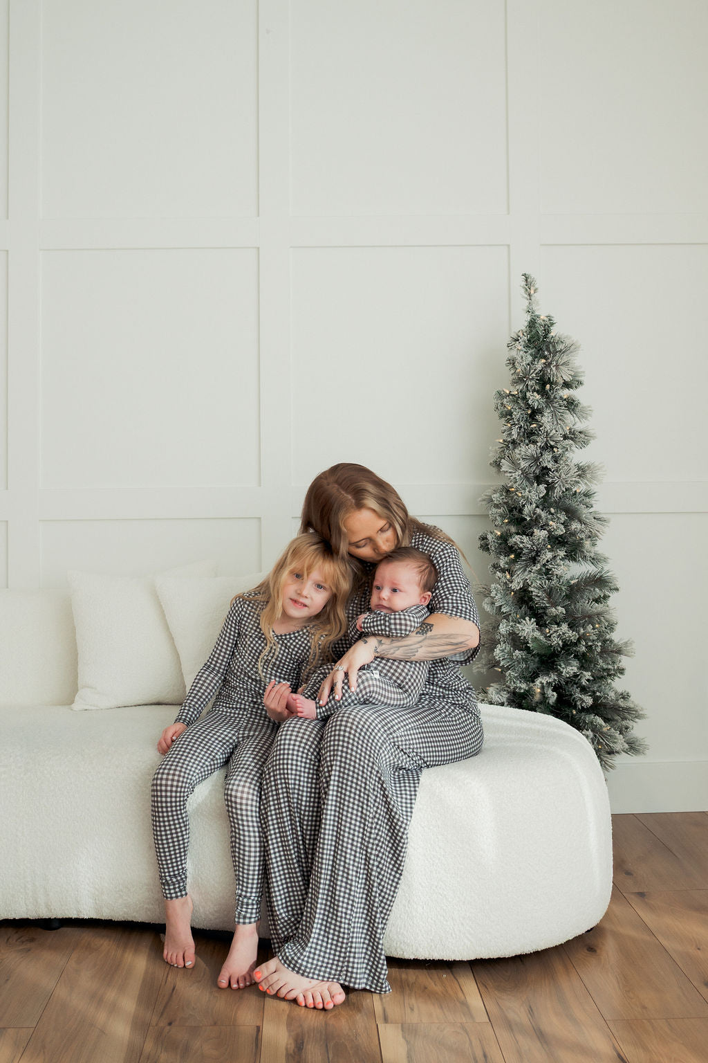 A woman in the forever french baby Women's Boxy Tee Lounge Set in Green Gingham hugs two children as they sit together on a white couch beside a frosted Christmas tree in a modern, minimal room.
