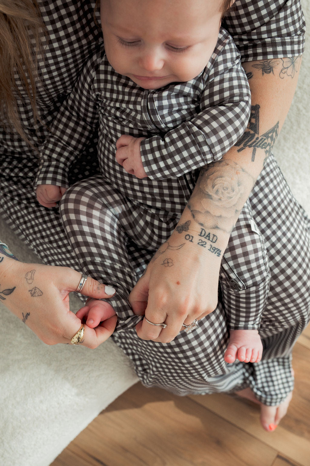 A baby in forever french baby's Bamboo Zip Pajamas in Green Gingham sits on an adult's lap, both dressed in matching outfits as the adult’s tattooed hands zip up the comfy, hypo-allergenic pajamas for style and comfort.