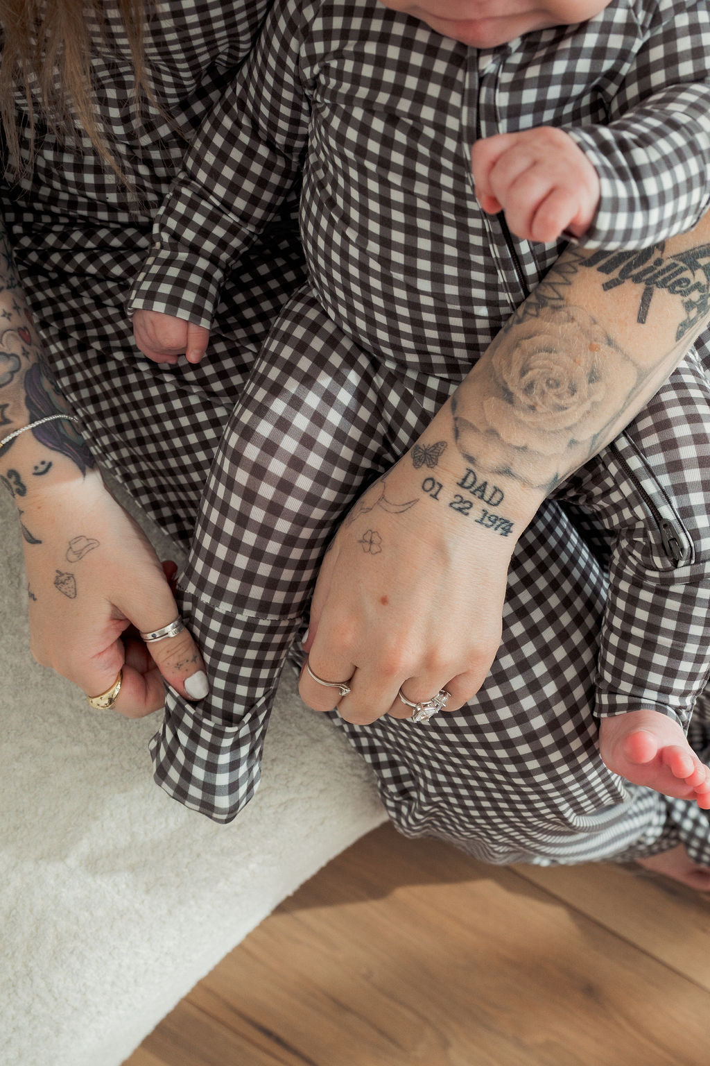 A tattooed adult and a baby, both in matching forever french baby Bamboo Zip Pajamas in Green Gingham, sit closely together. The adult’s hand with rings and tattoos gently rests on the baby's leg.