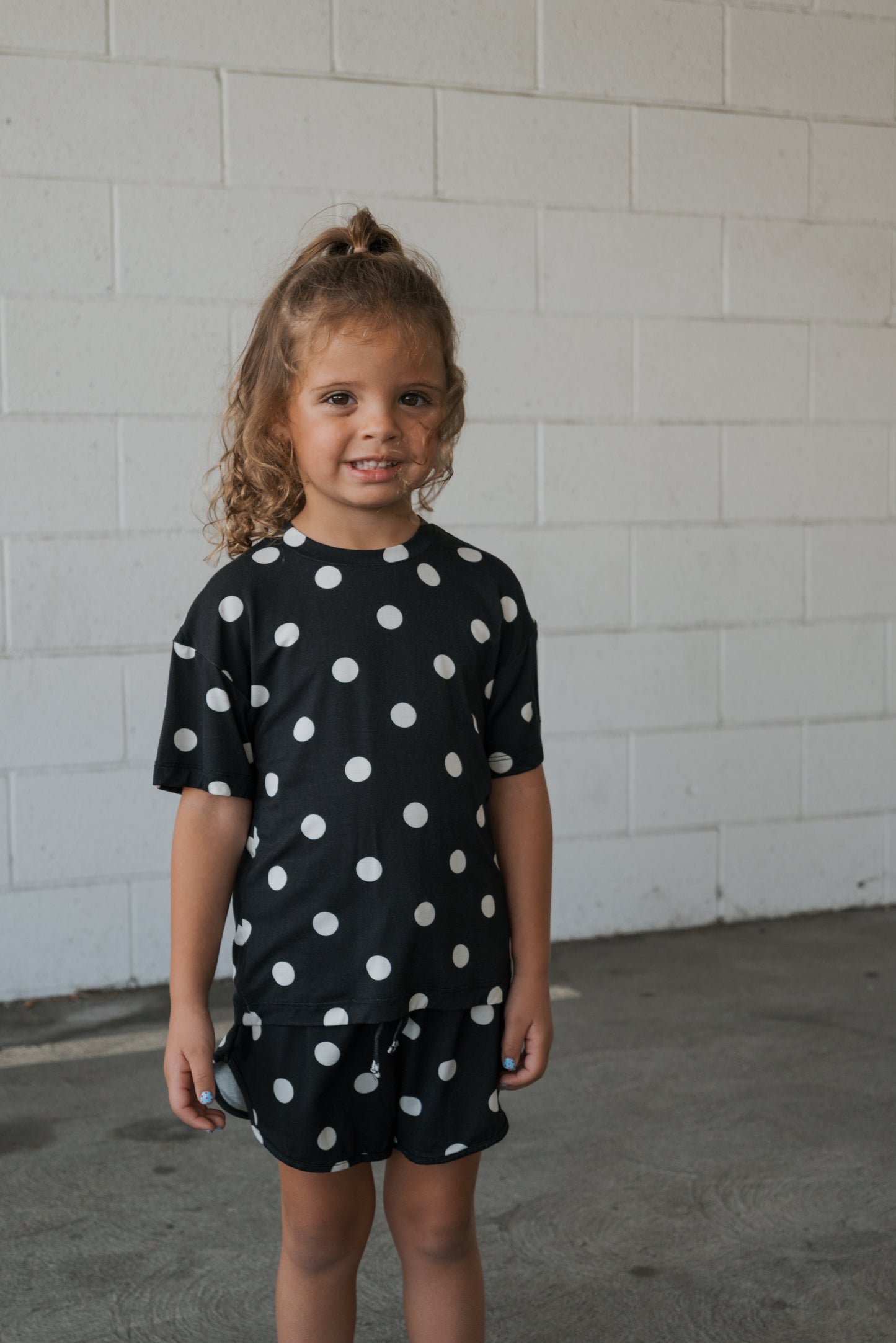 A young child with curly hair, dressed in the forever french baby Girl’s Bamboo Short Set in classic black and white polka dot, stands smiling against a white brick wall—perfectly highlighting the gentle, hypo-allergenic comfort of bamboo clothing for sensitive skin.