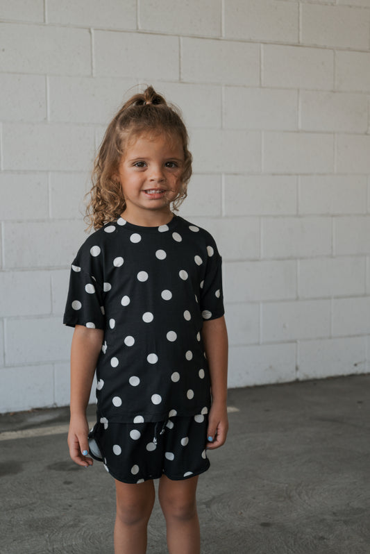 A young child with curly hair, dressed in the forever french baby Girl’s Bamboo Short Set in classic black and white polka dot, stands smiling against a white brick wall—perfectly highlighting the gentle, hypo-allergenic comfort of bamboo clothing for sensitive skin.