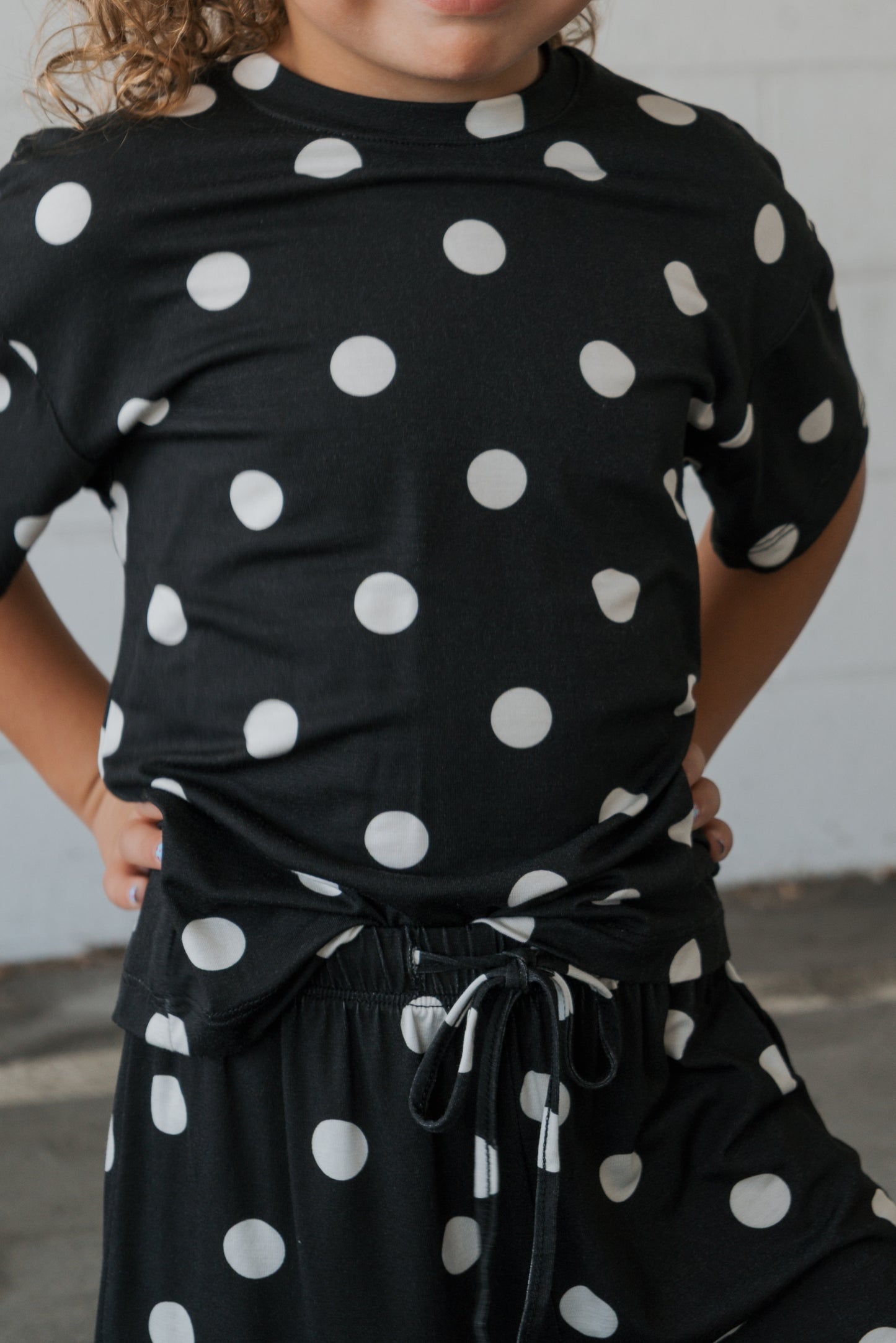 A child stands against a gray wall with hands on hips, dressed in the forever french baby Girl’s Bamboo Short Set | B&W Polka Dot—an outfit crafted from hypo-allergenic bamboo featuring bold white polka dots on black. The child’s face is partially out of frame, keeping attention on the stylish set.