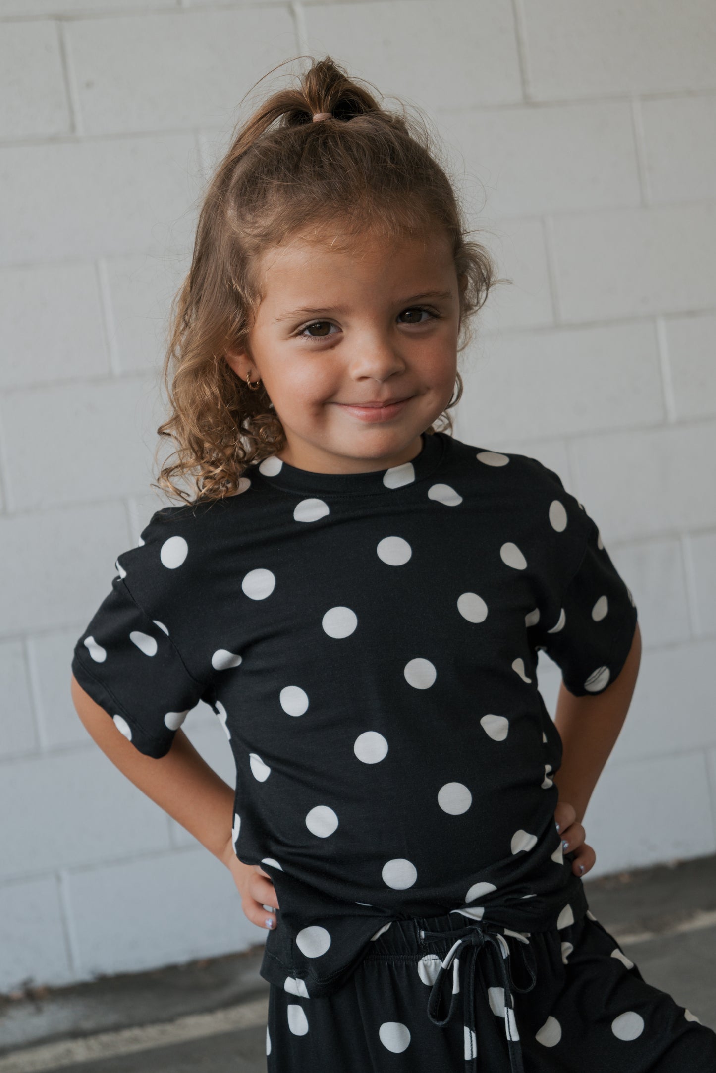 A young child smiles confidently, hands on hips, wearing the Girl’s Bamboo Short Set | B&W Polka Dot by forever french baby in front of a white brick wall. This stylish set is made from hypo-allergenic bamboo, making it ideal for sensitive skin.