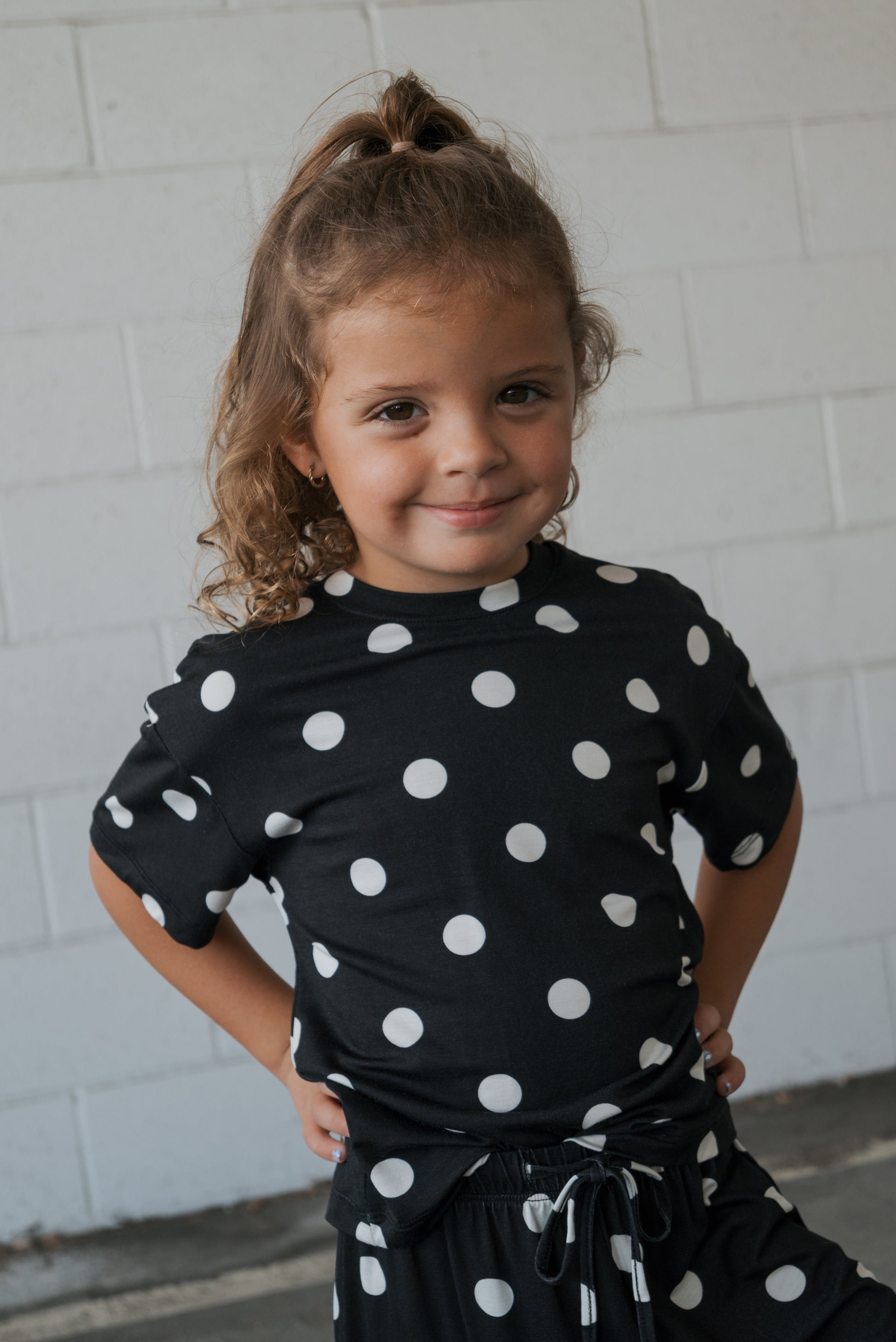 A young child smiles confidently, hands on hips, wearing the Girl’s Bamboo Short Set | B&W Polka Dot by forever french baby in front of a white brick wall. This stylish set is made from hypo-allergenic bamboo, making it ideal for sensitive skin.