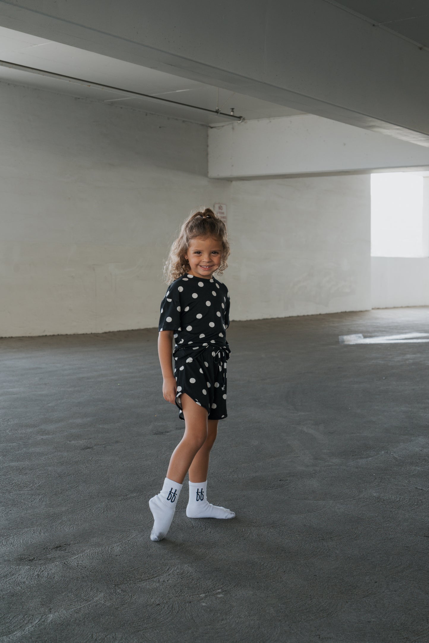 A young child stands and smiles in a spacious, empty concrete room with white walls and large windows, dressed in the forever french baby Girl’s Bamboo Short Set | B&W Polka Dot and white socks. The hypo-allergenic bamboo fabric ensures comfort while they play.