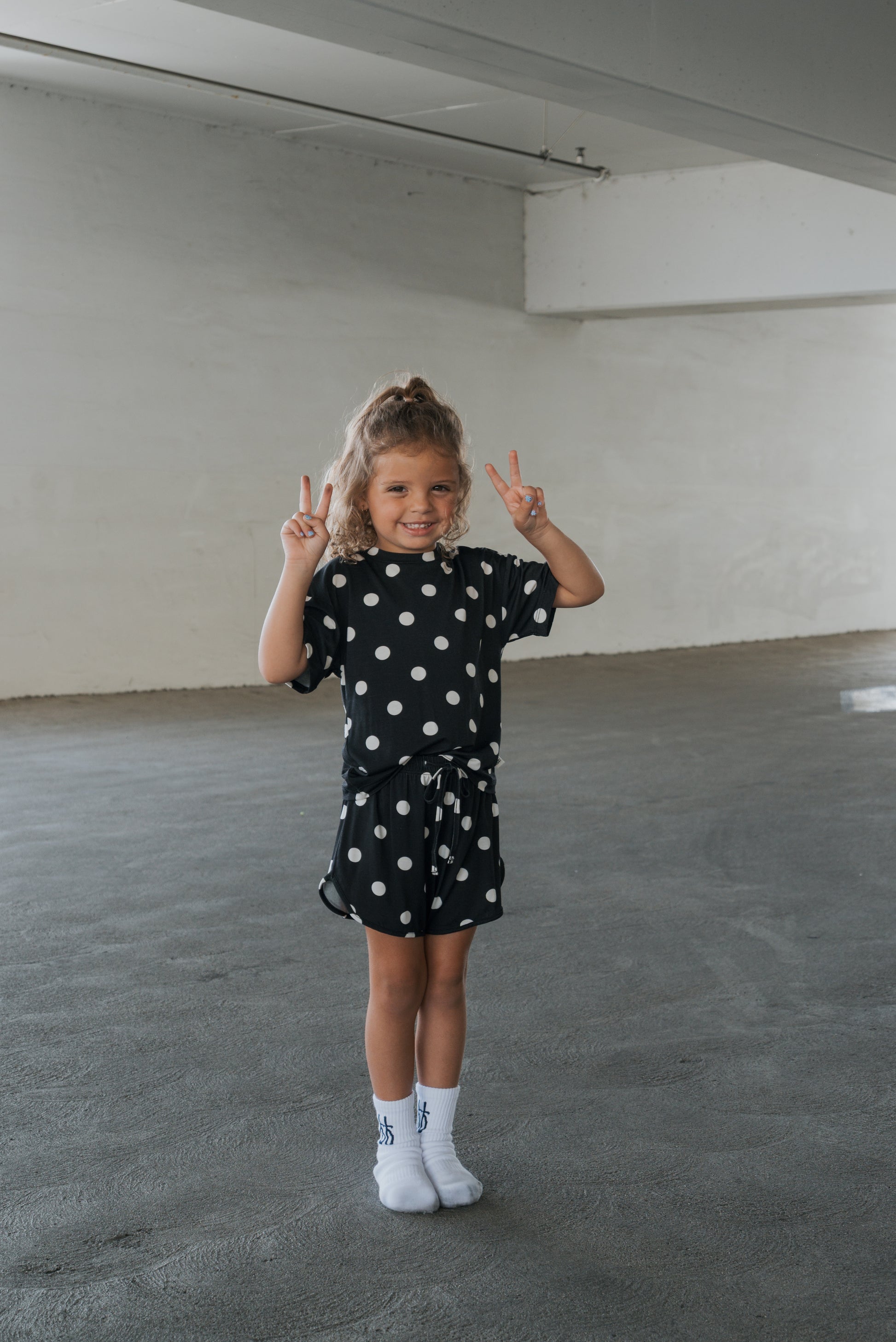 A young child stands indoors on a concrete floor, smiling brightly and flashing two peace signs with both hands while wearing the forever french baby Girl’s Bamboo Short Set | B&W Polka Dot, radiating happiness and comfort in her soft, hypo-allergenic bamboo outfit.