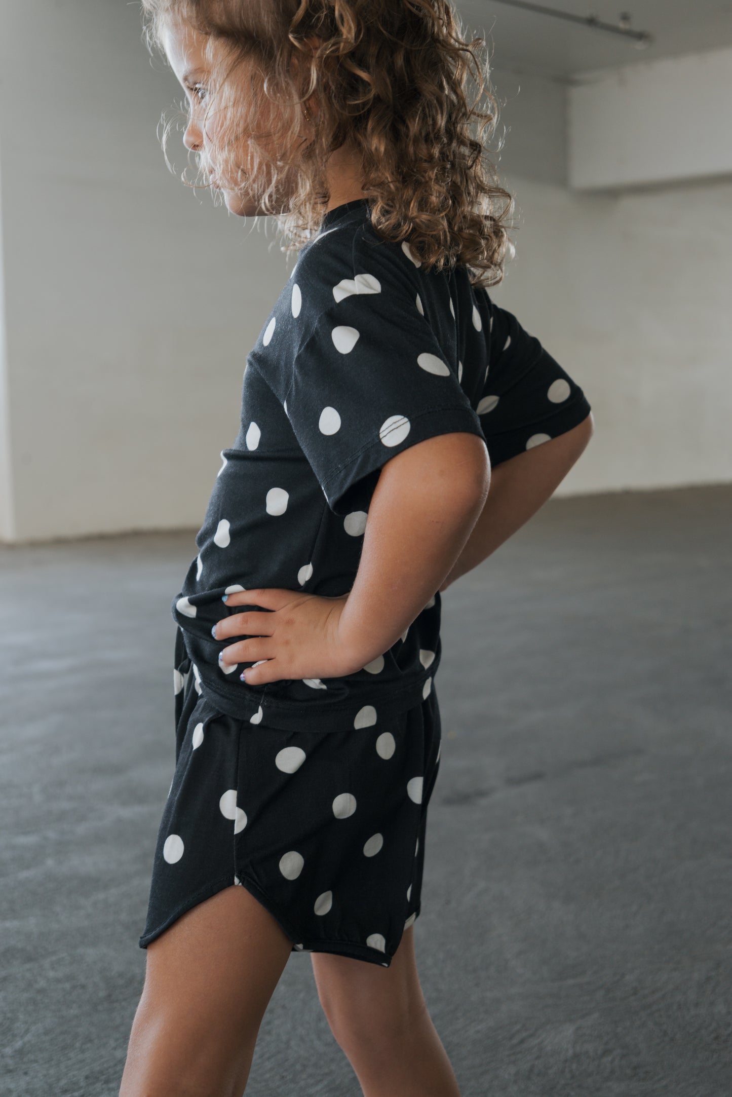 A young child with curly hair stands sideways in a room, hands on hips, wearing the forever french baby Girl’s Bamboo Short Set in B&W Polka Dot. This hypo-allergenic set offers both comfort and style.