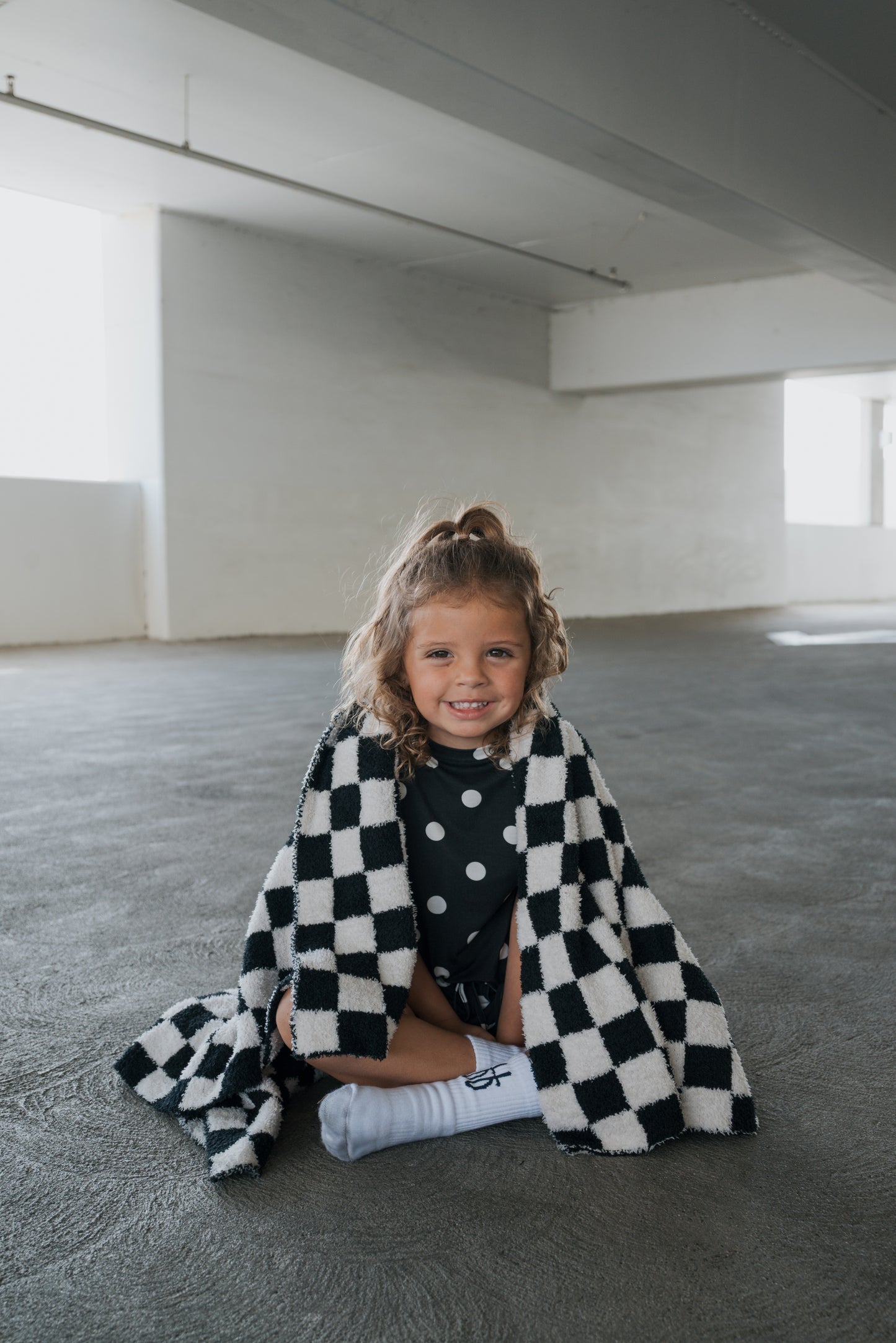 A young child with curly hair sits on a concrete floor, smiling and wrapped in the forever french baby Plush Blanket | B&W Micro Check, wearing a polka dot outfit and white socks in a bright, empty room.