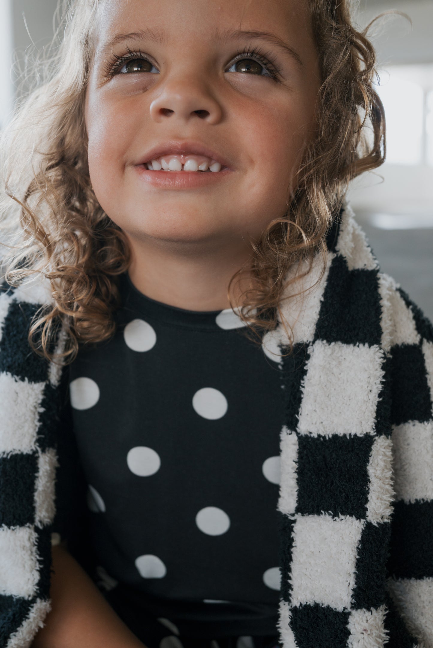 A young child with curly hair smiles while wearing the forever french baby Girl’s Bamboo Short Set in black-and-white polka dot, draped with a checkered blanket over their shoulders, highlighting the comfort of hypo-allergenic bamboo clothing.