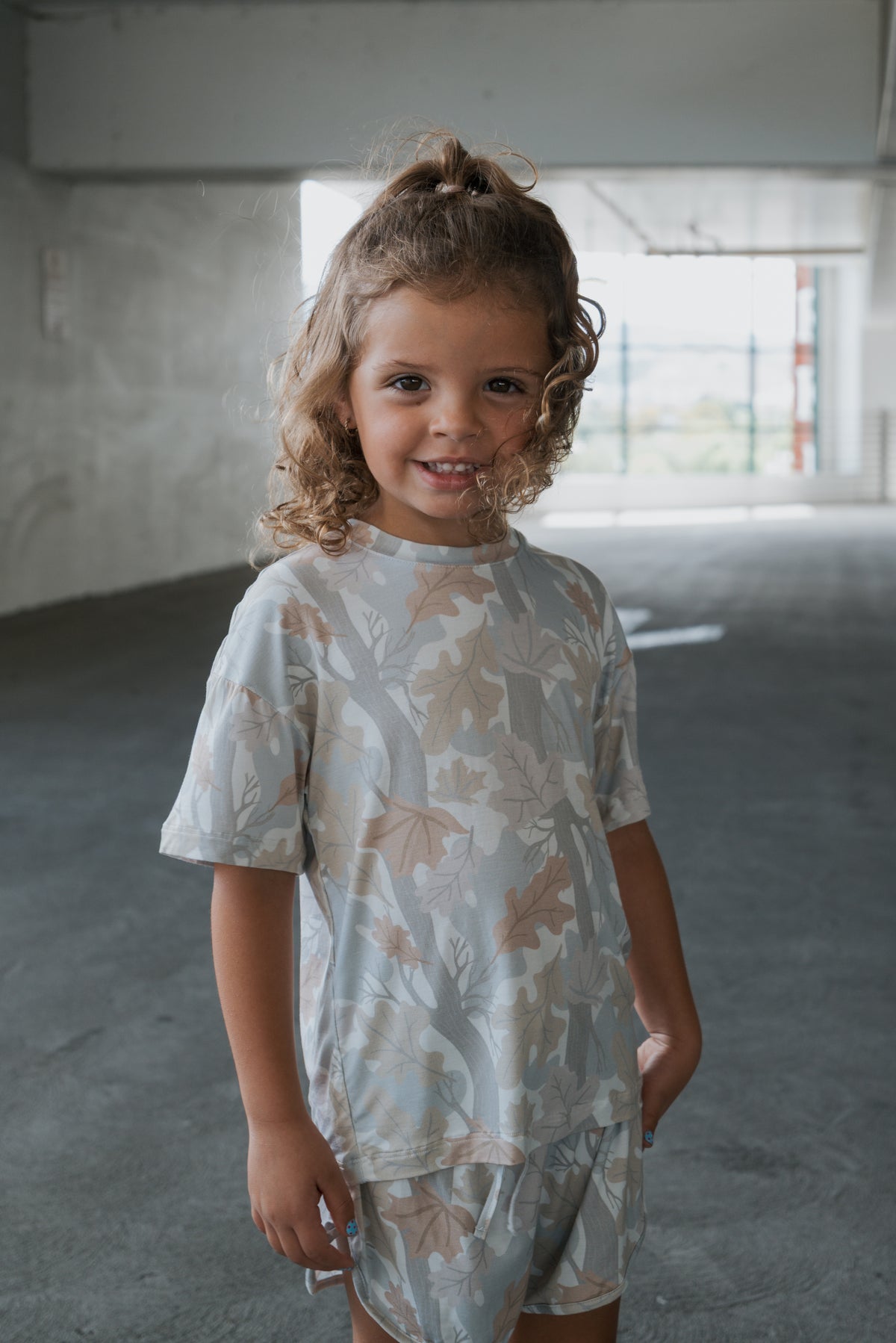 A young child with curly hair, wearing the forever french baby Girl’s Bamboo Short Set in Woodland, smiles while standing in a bright, empty indoor space.