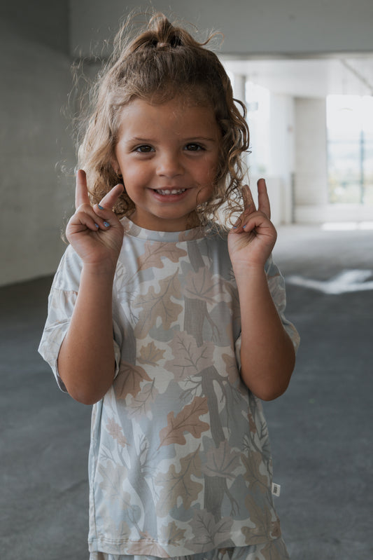 A young child with curly hair smiles and holds up both hands with fingers crossed, standing indoors in a softly lit, modern space while wearing the forever french baby Girl’s Bamboo Short Set | Woodland—perfect for little ones who need hypo-allergenic children’s clothing.