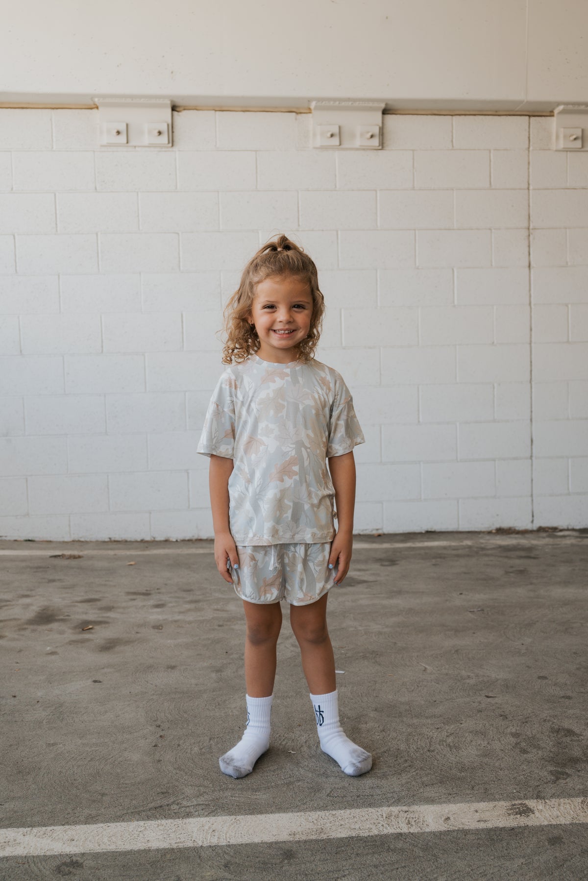A young child with curly hair stands smiling in the forever french baby Girl’s Bamboo Short Set | Woodland and white socks on a concrete floor, with a white brick wall in the background.