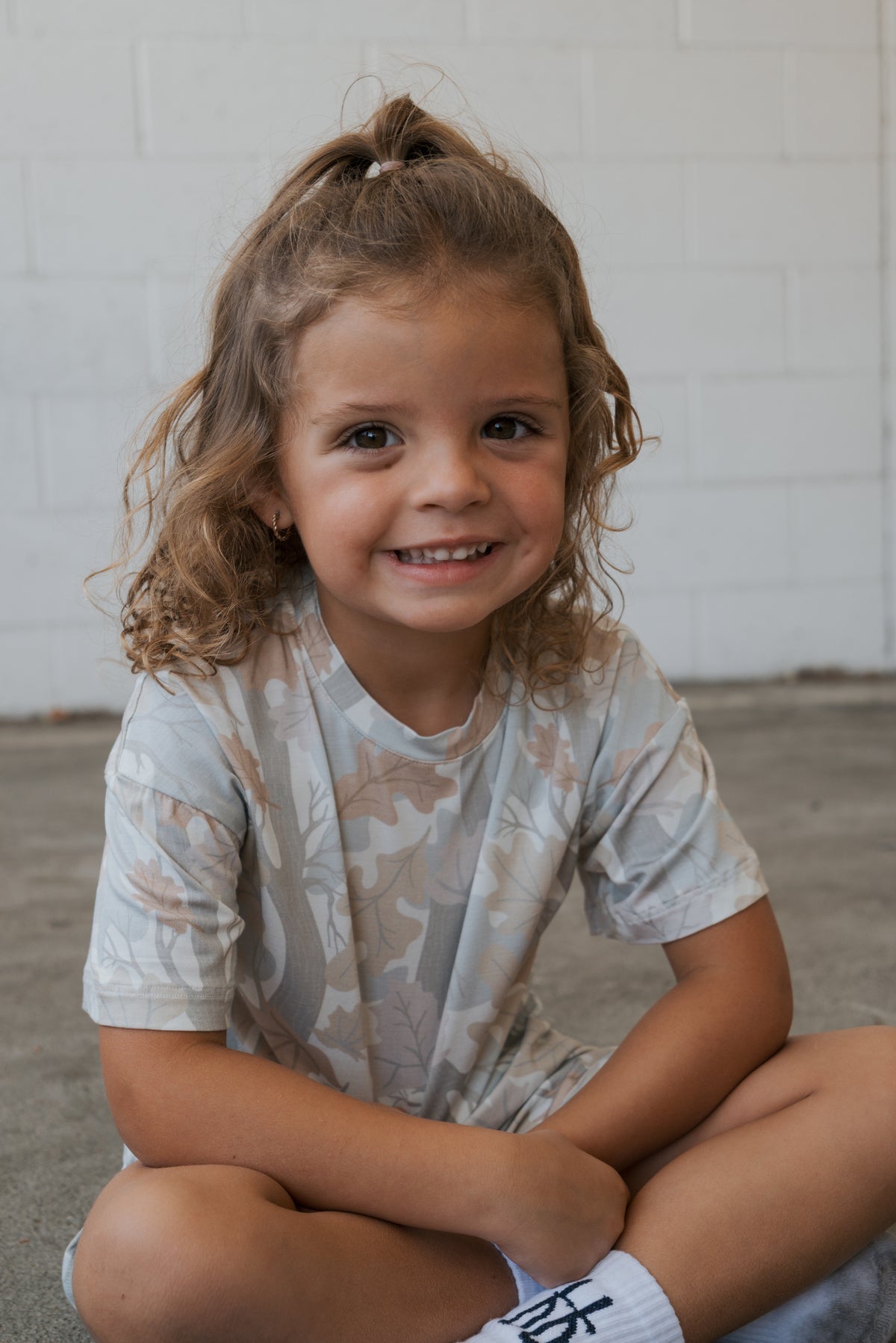 A young child with curly hair sits cross-legged on the ground, smiling, wearing the forever french baby Girl’s Bamboo Short Set | Woodland and white socks. The background is a white brick wall.