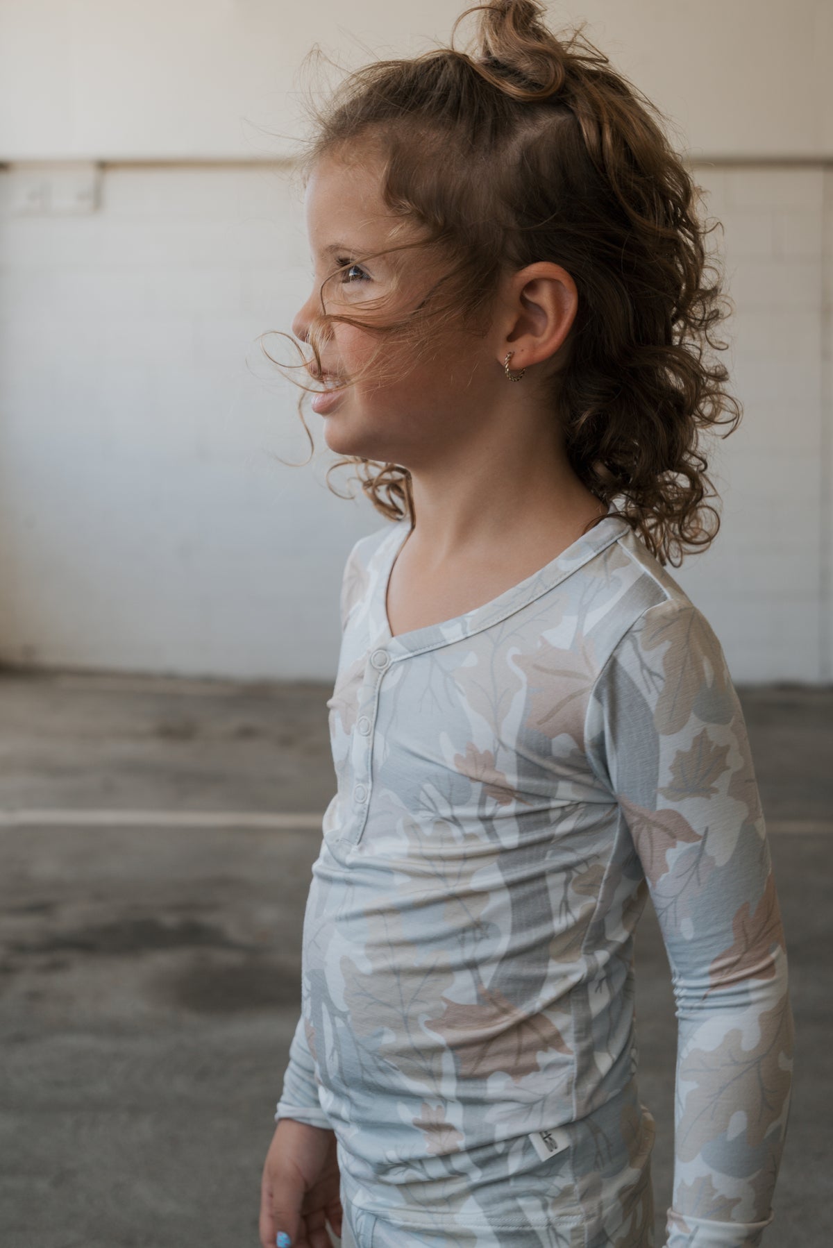 A young child with curly hair, wearing the forever french baby Bamboo Two Piece Pajamas in the Woodland print—a long-sleeve, light-colored pajama shirt with a leafy pattern—stands indoors, smiling and looking to the left.