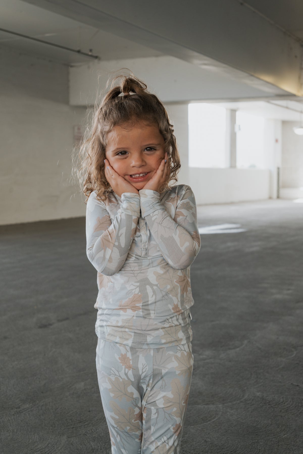 A young girl with curly hair smiles and holds her face with both hands as she stands in a light-filled indoor space, wearing the forever french baby Bamboo Two Piece Pajamas in the Woodland pattern.