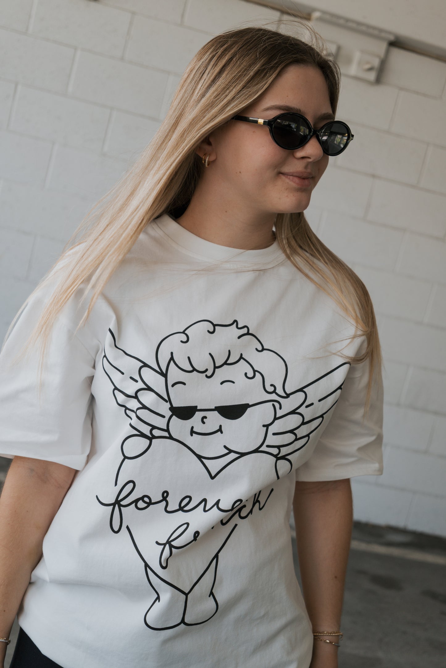 A woman wearing sunglasses and a white Adult Cotton Tee from forever french baby, featuring the Big Baby cartoon angel in sunglasses with the word “forever,” stands in front of a white brick wall, showcasing this unisex everyday cotton t-shirt.