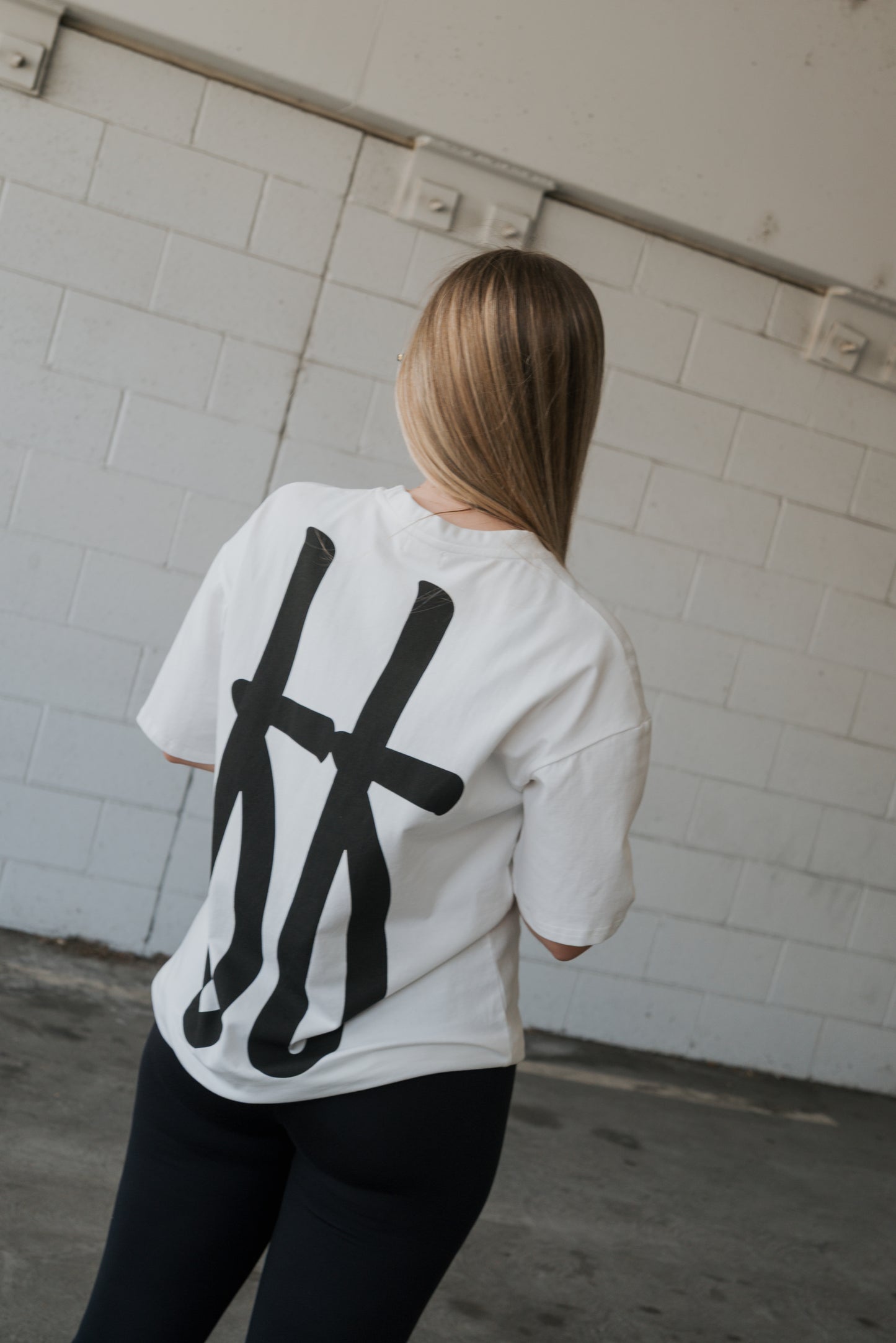 A person with long blonde hair, wearing the forever french baby Adult Cotton Tee | Big Baby—a unisex cotton t-shirt showcasing a bold black graphic design on the back—stands indoors facing a white brick wall, embodying effortless everyday style.