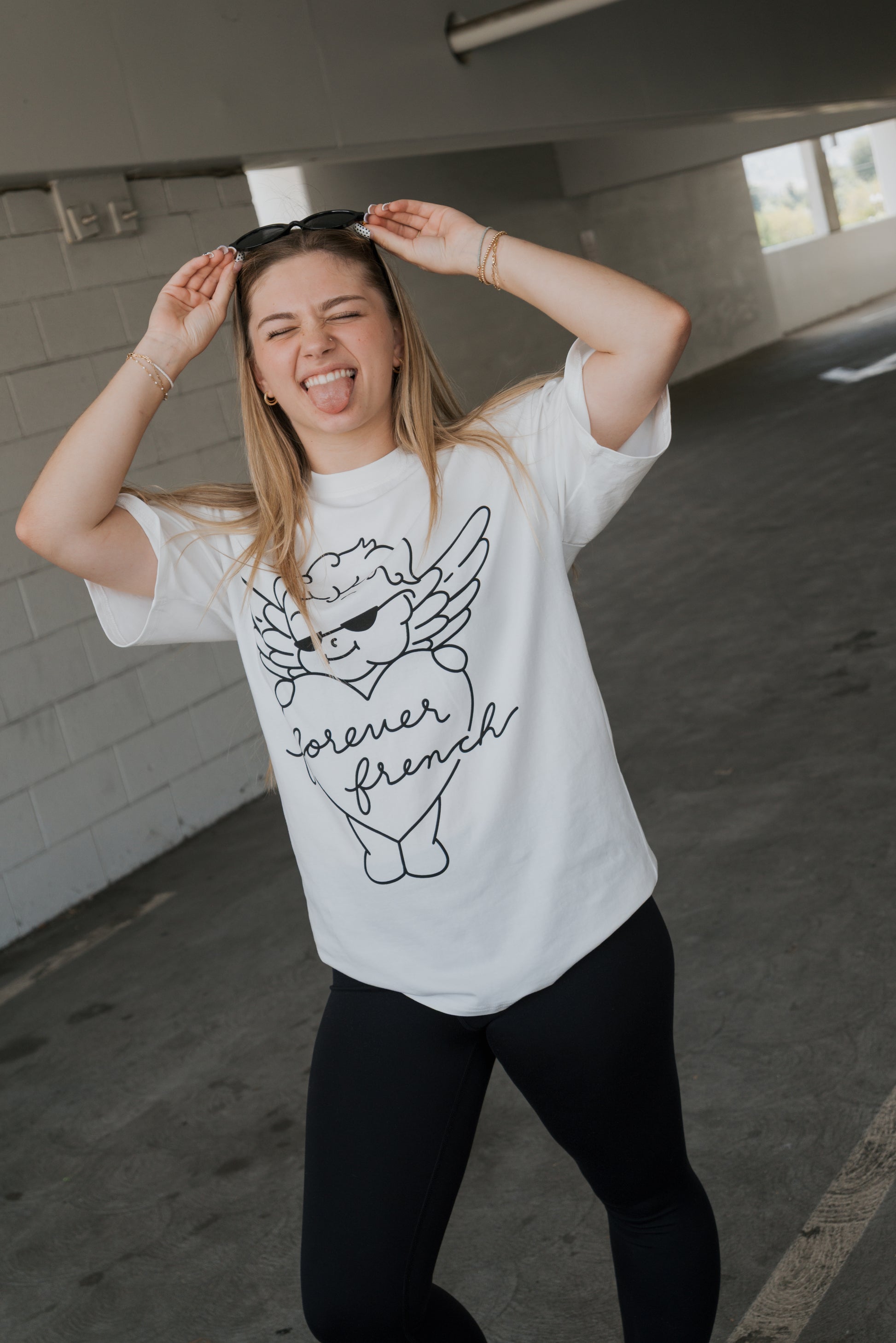 A young woman wearing a forever french baby Adult Cotton Tee | Big Baby and black leggings stands in a parking garage, smiling and sticking out her tongue while lifting her sunglasses with both hands—showcasing effortless style perfect for everyday wear.