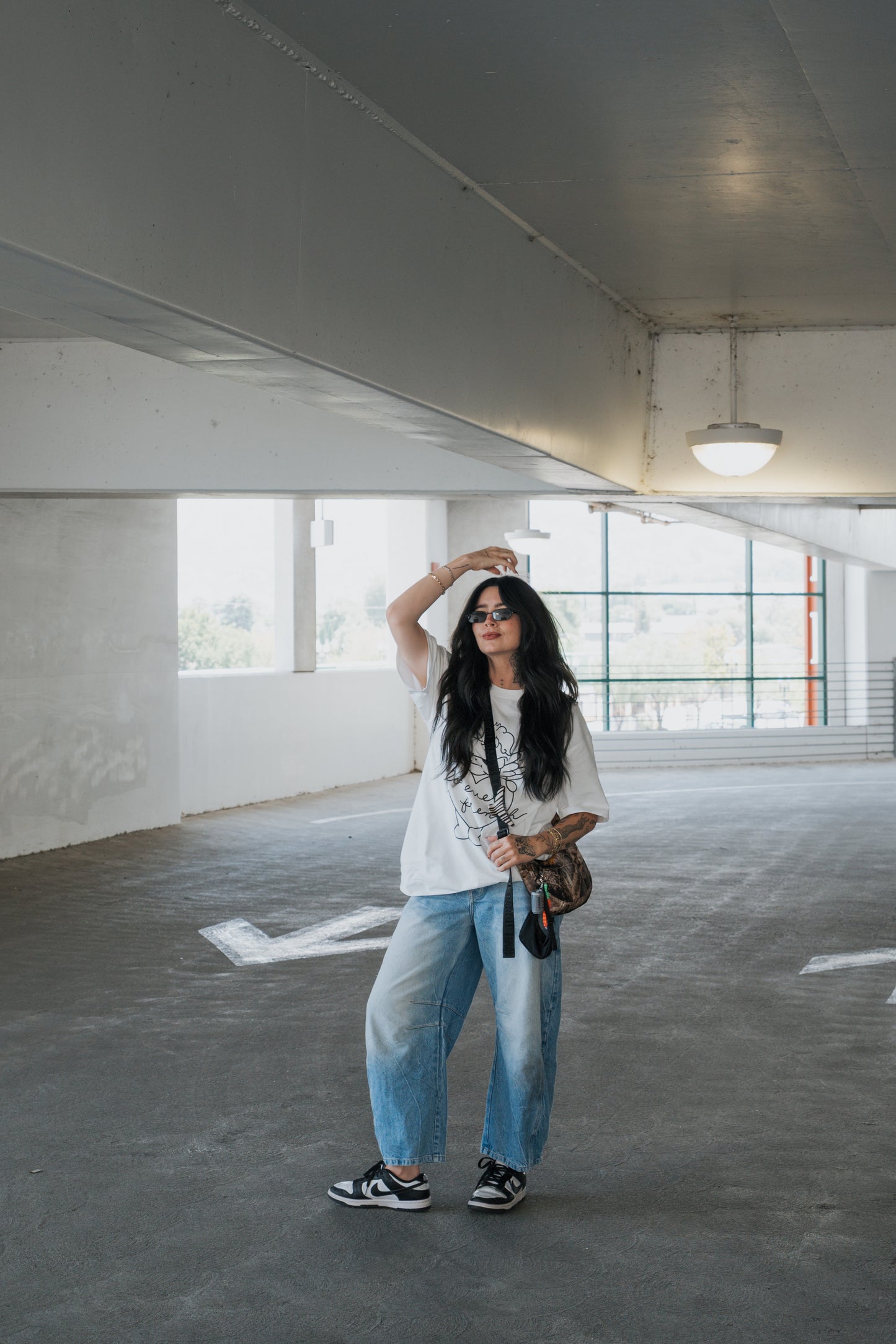 A woman stands in a parking garage, wearing sunglasses and the forever french baby Adult Cotton Tee | Big Baby—an effortlessly stylish unisex cotton t-shirt ideal for everyday wear—paired with wide-legged jeans and sneakers. She holds a bag and keys, one hand raised to her head, exuding a relaxed and fashionable vibe.