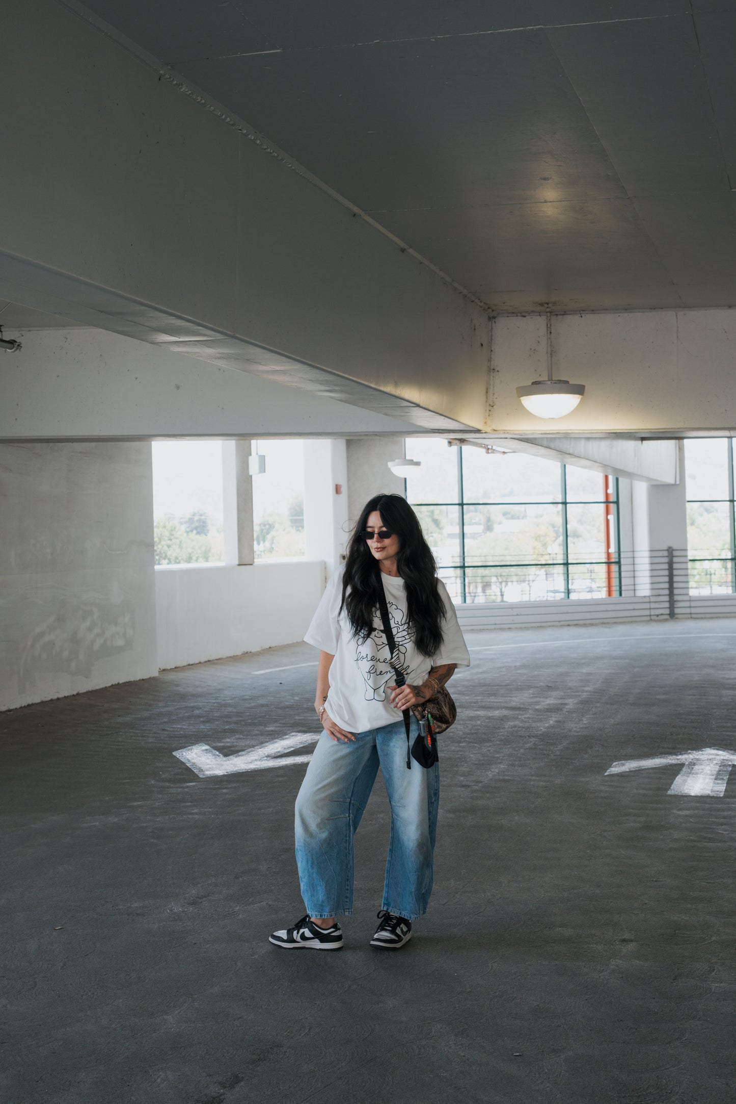 A person with long dark hair, sunglasses, a white Adult Cotton Tee by forever french baby, loose blue jeans, and sneakers stands in a parking garage holding a small bag, framed by large windows in the background—an effortlessly cool take on everyday wear.