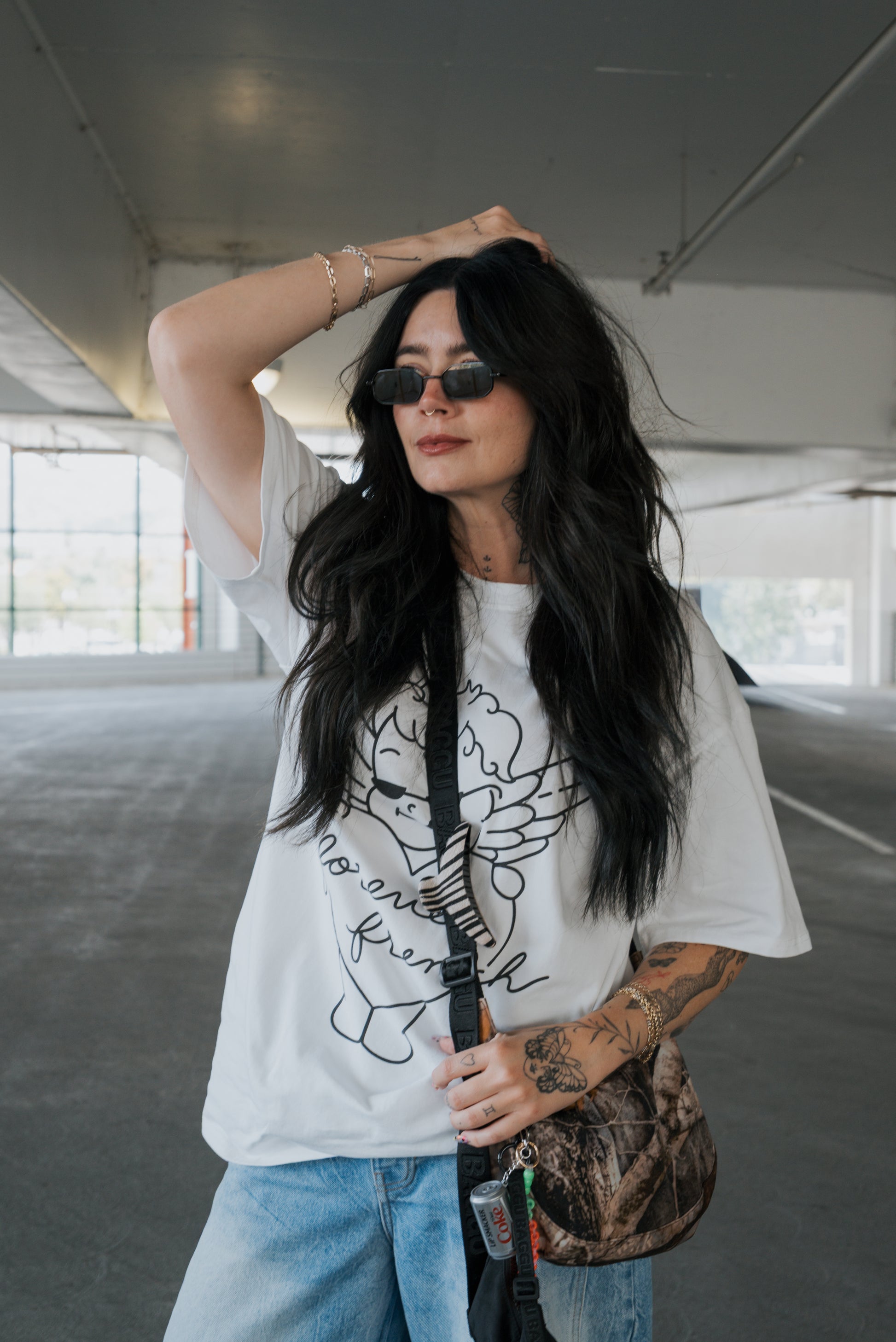 A woman with long dark hair, sunglasses, and tattoos poses in a parking garage wearing the Adult Cotton Tee | Big Baby by forever french baby—a crisp white t-shirt perfect for everyday wear—paired with light blue jeans, a crossbody bag, and her hand resting on her head.