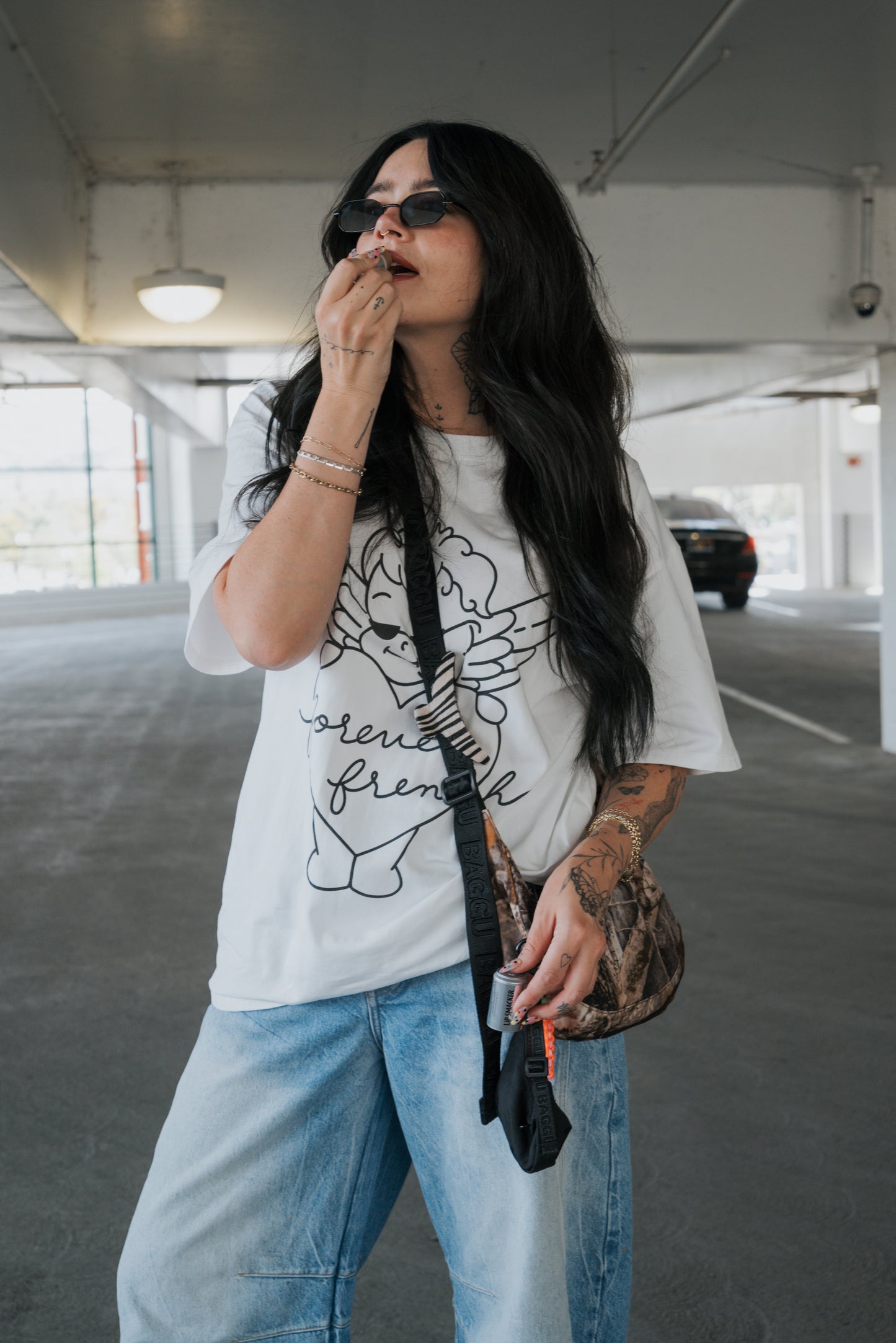 A woman wearing sunglasses, light jeans, and the forever french baby Adult Cotton Tee | Big Baby applies lip balm in a parking garage, holding a brown bag with a strap over her shoulder. This versatile unisex cotton tee from forever french baby is perfect for everyday wear.