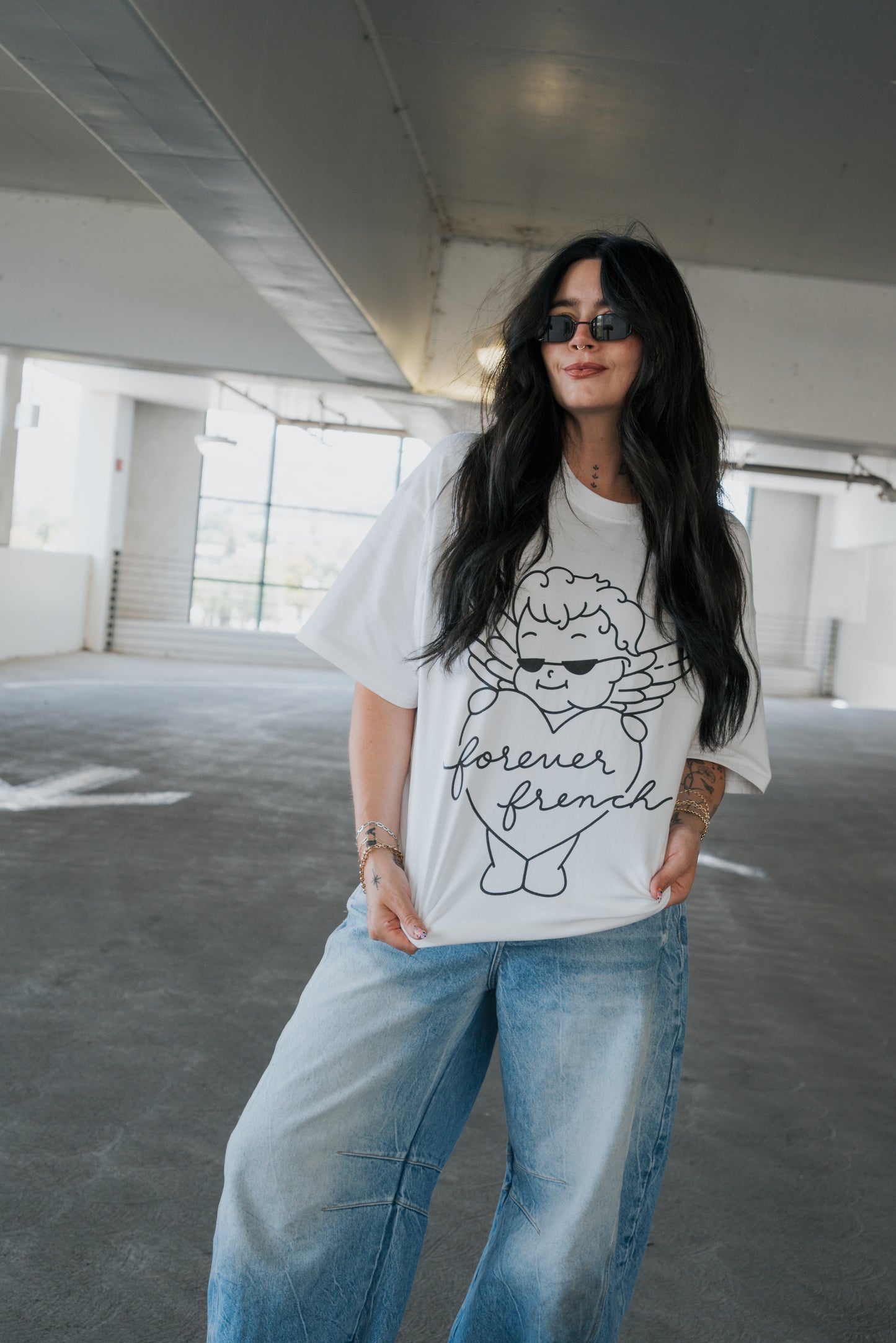 A woman with long dark hair, wearing sunglasses, a white “Adult Cotton Tee | Big Baby” from forever french baby featuring a cartoon graphic, and loose blue jeans stands in a bright, empty parking garage, smiling slightly with one hand in her pocket—perfectly capturing the effortless vibe of everyday wear.