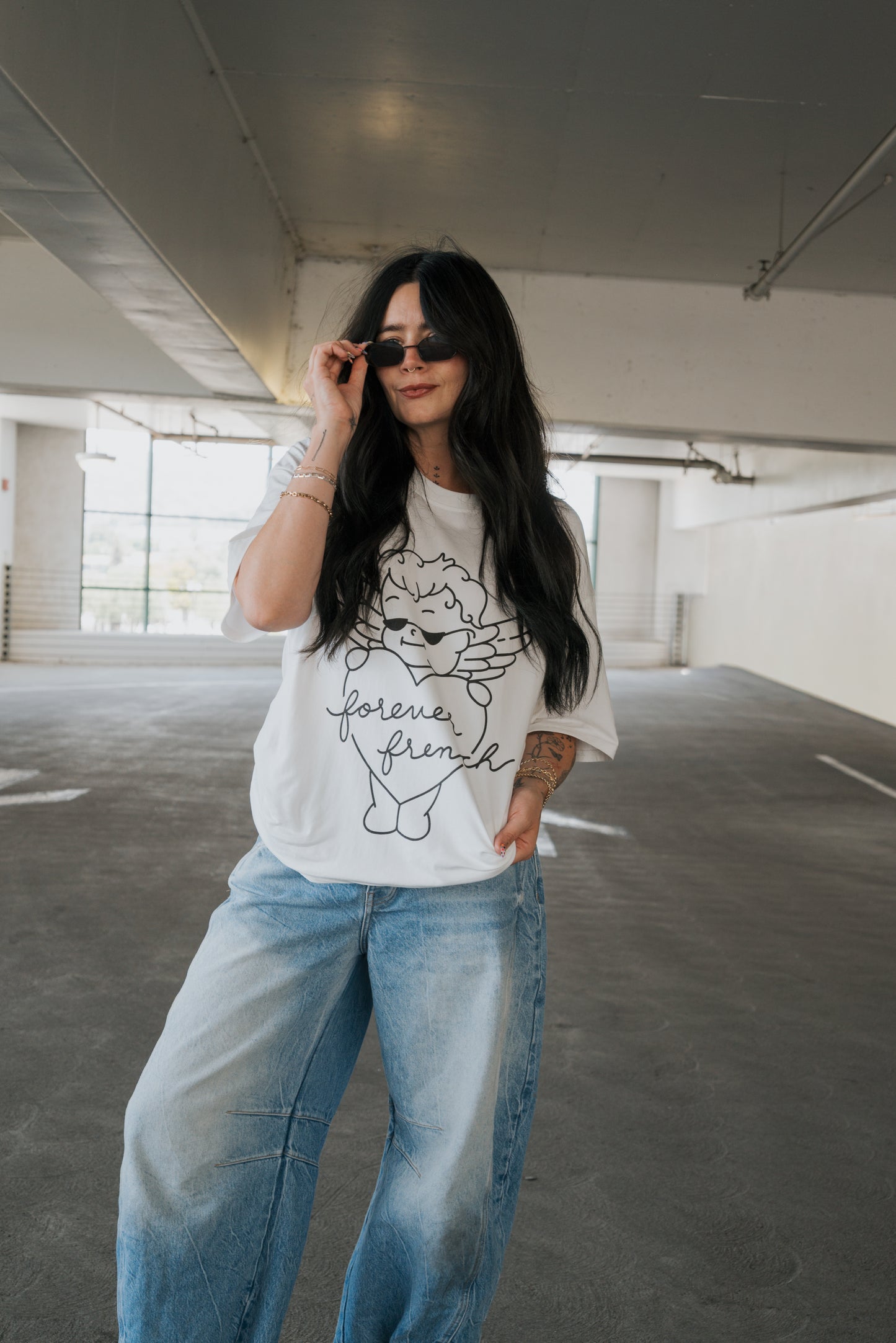 A woman with long dark hair, wearing sunglasses, a white “forever french baby” Adult Cotton Tee | Big Baby, and loose blue jeans stands in a parking garage, smiling confidently with one hand in her pocket—perfect everyday wear for effortless style.