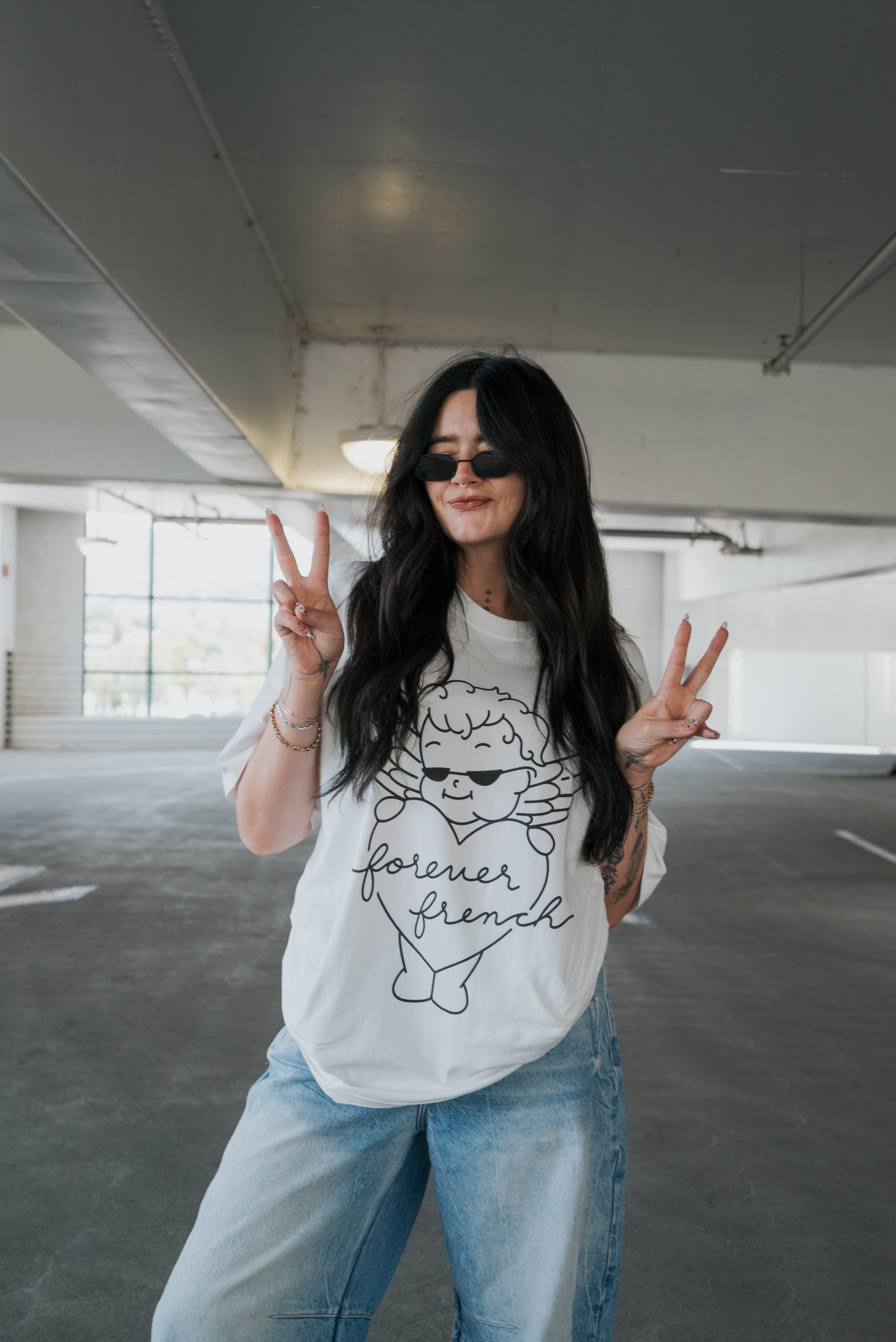 A woman with long dark hair, wearing sunglasses, jeans, and the “Adult Cotton Tee” by forever french baby featuring a white “forever friend” graphic, stands in a parking garage making peace signs and smiling—showcasing this unisex everyday cotton t-shirt.